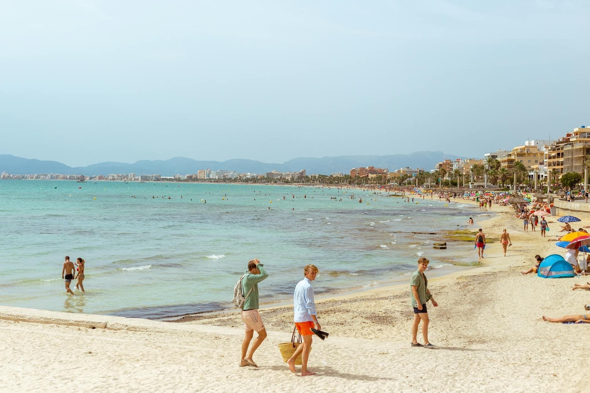 Menschen spazieren und entspannen an einem Sandstrand auf Mallorca mit türkisblauem Wasser, Sonnenschirmen und lebhafter Sommeratmosphäre im Juli oder August.