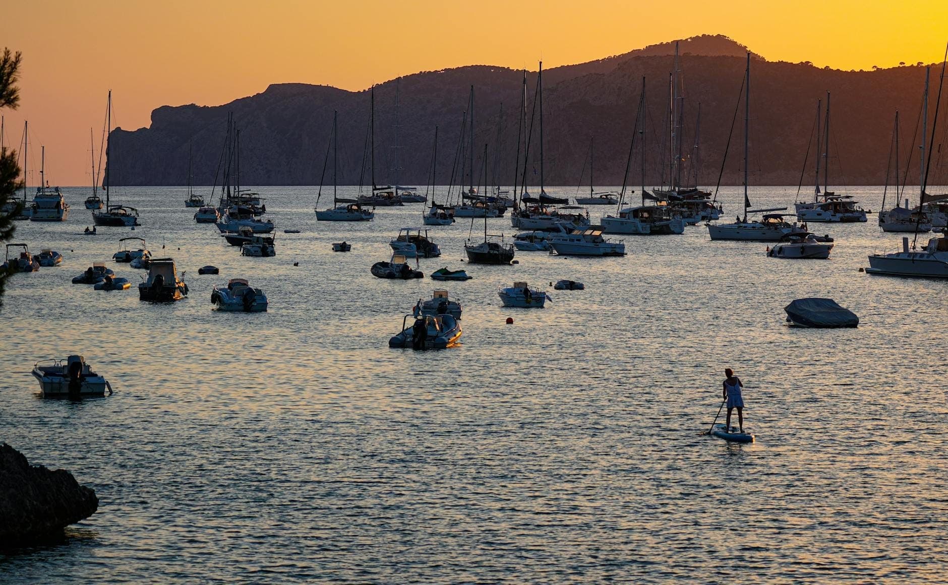Barche e yacht ancorati al tramonto sulle calme acque del Mediterraneo con colline sullo sfondo, che evocano l'atmosfera delle feste in barca serali a Mallorca.