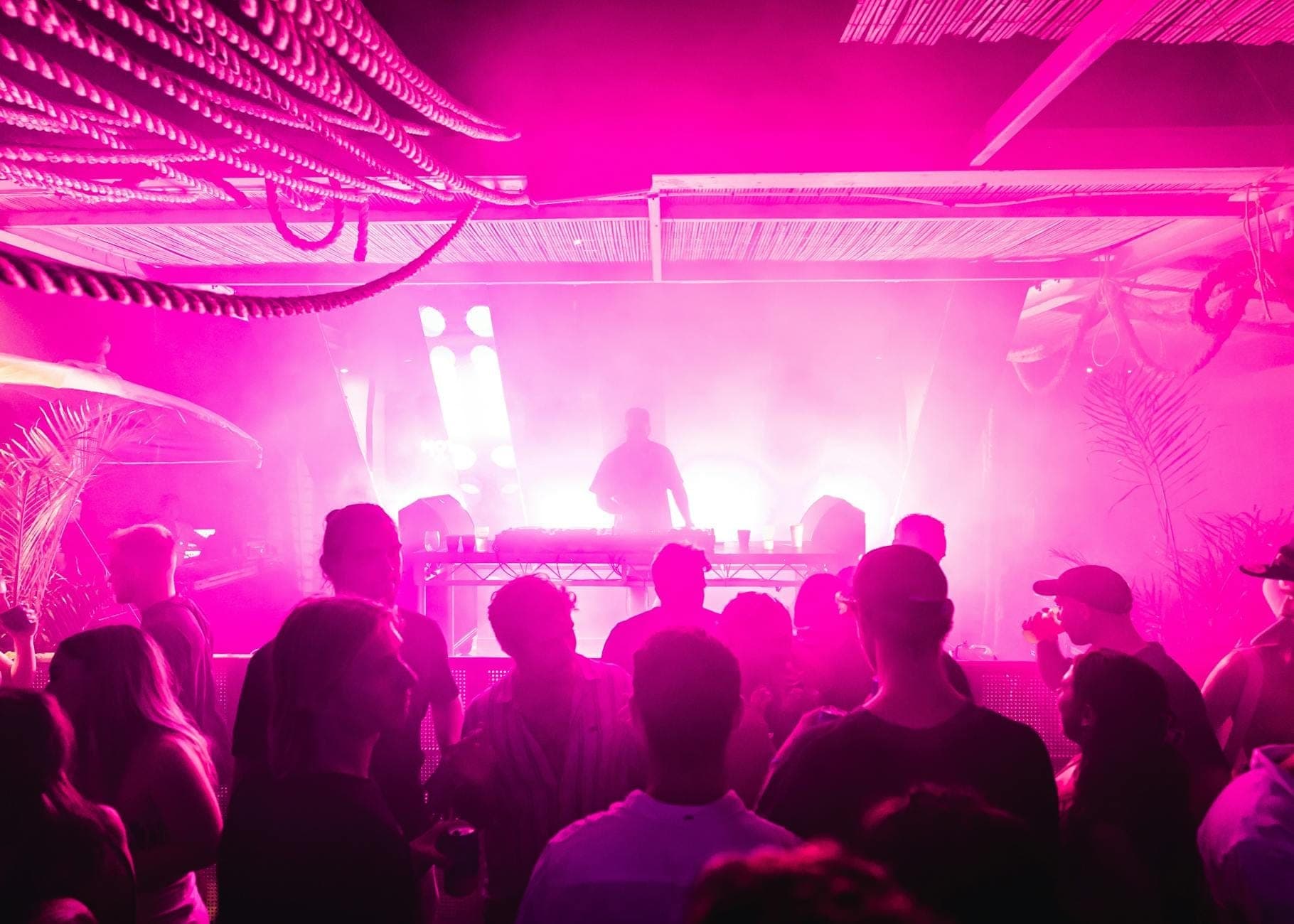 Menschenmenge auf einer lebhaften Clubnacht oder Strandparty auf Mallorca mit pink-violettem Licht und einem DJ auf der Bühne.