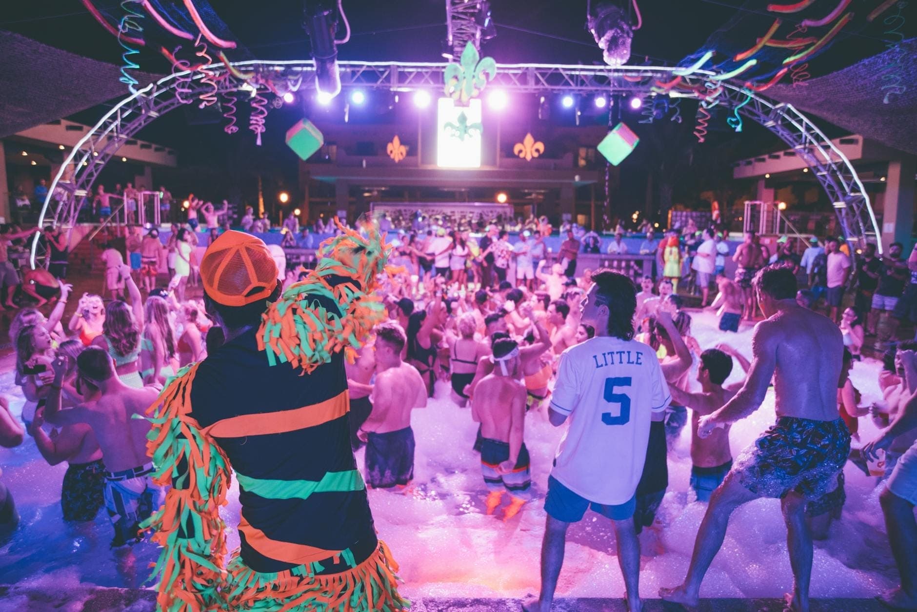 Grande folla a una festa della schiuma notturna all'aperto con luci del palco colorate e un'atmosfera vivace tipica dei club dei resort mediterranei.