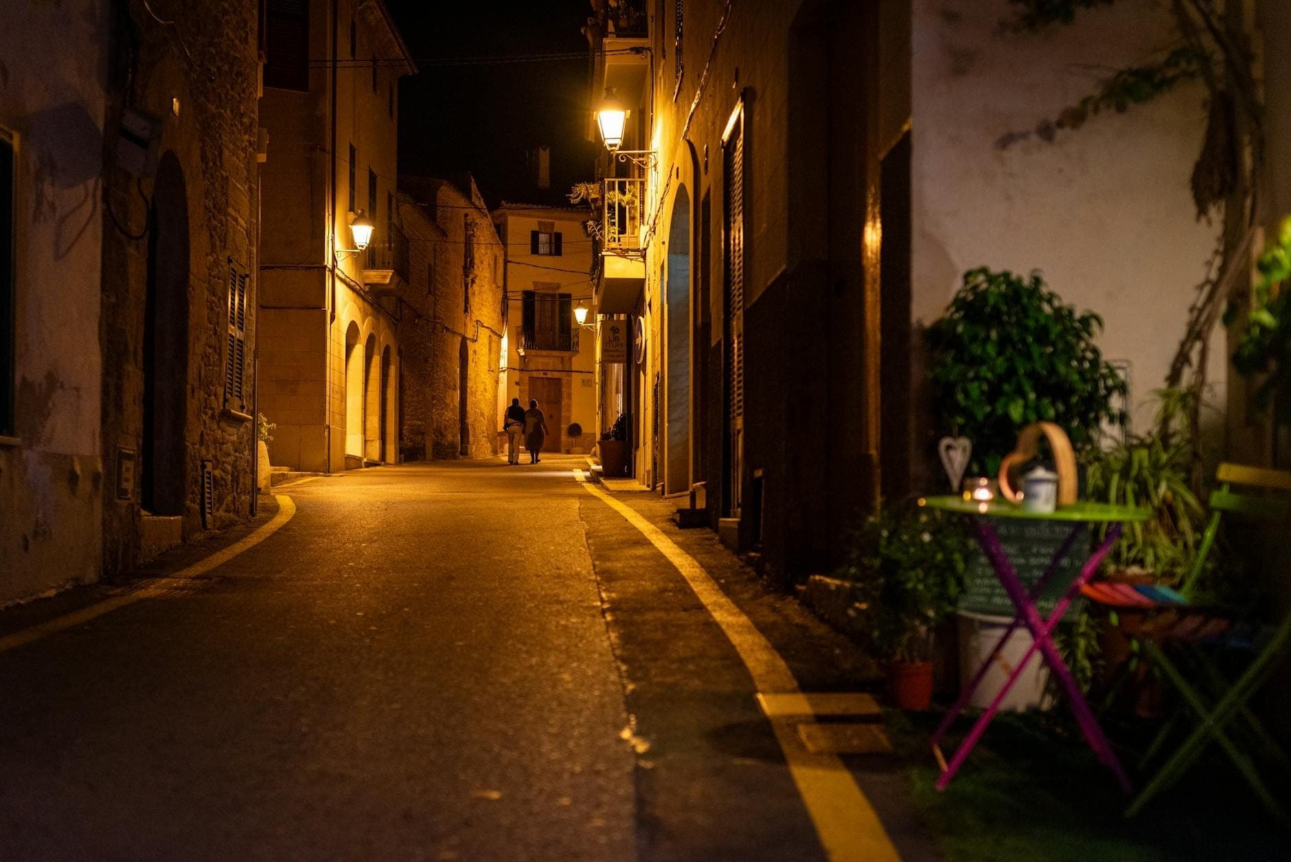 Una strada stretta e calorosamente illuminata di notte con storici edifici in pietra, lampioni e un accogliente tavolo da caffè con sedie lungo il muro.