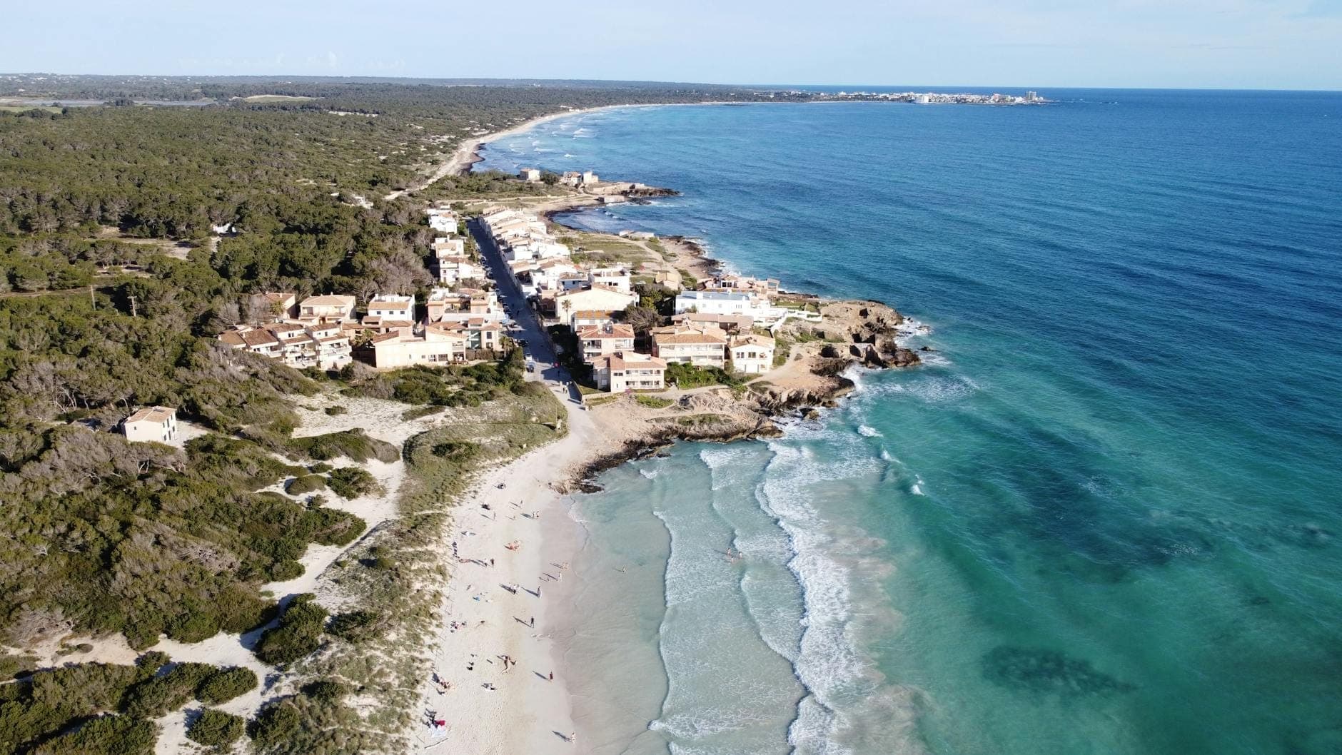 Vista aerea di una spiaggia di Maiorca con acque turchesi, villaggio costiero e foreste verdi lungo la riva in una giornata di sole.