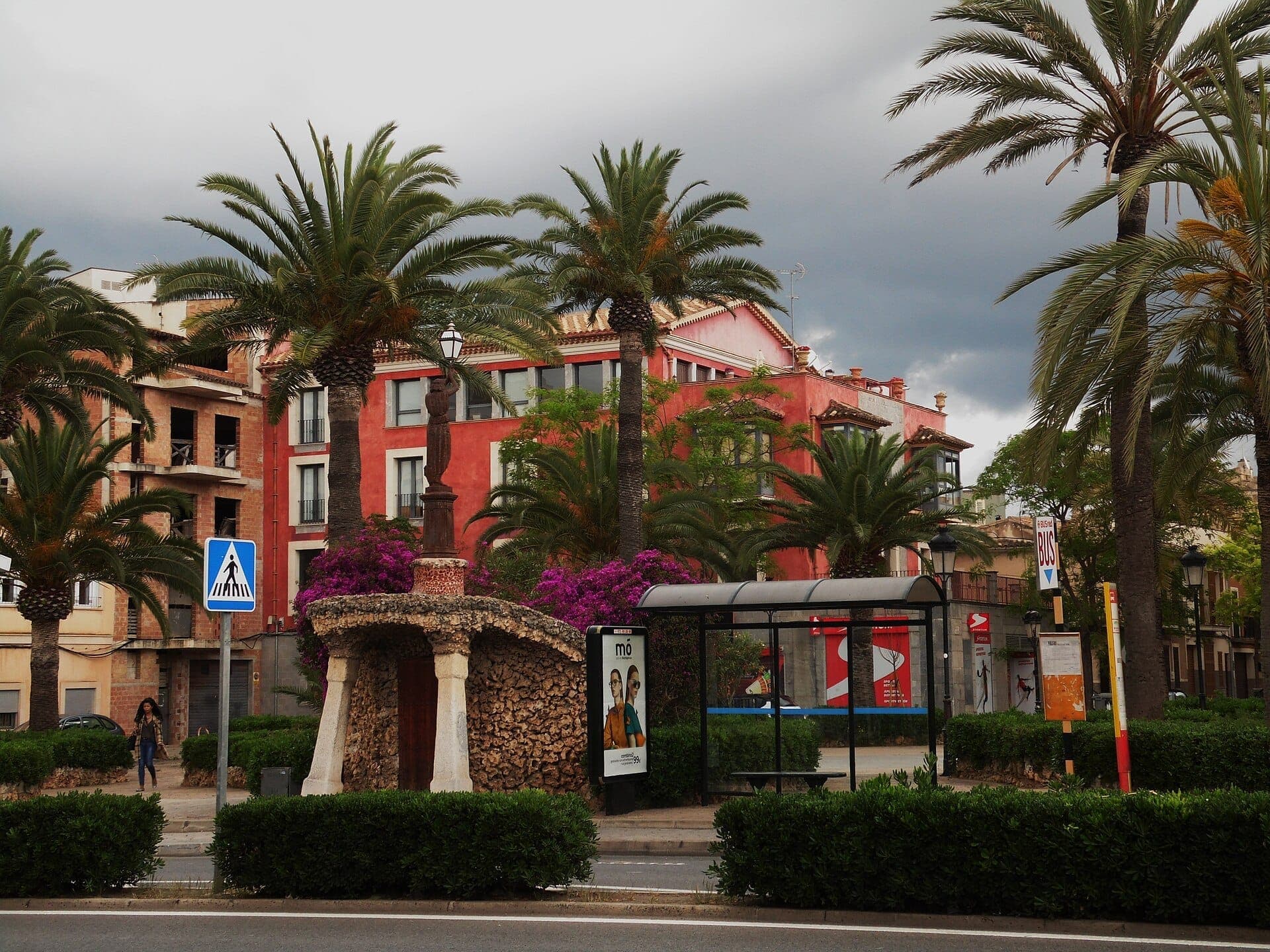 Straßenansicht in Manacor mit Palmen, einem Steinmonument, rosa Gebäuden und einer überdachten Bushaltestelle unter bewölktem Himmel.