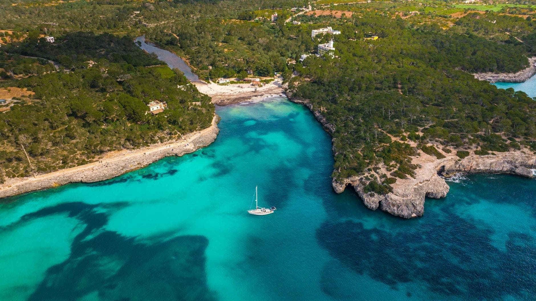 Una splendida vista aerea di una baia naturale con acque turchesi circondata da foresta verde e costa rocciosa, con una barca a vela ancorata nella baia.