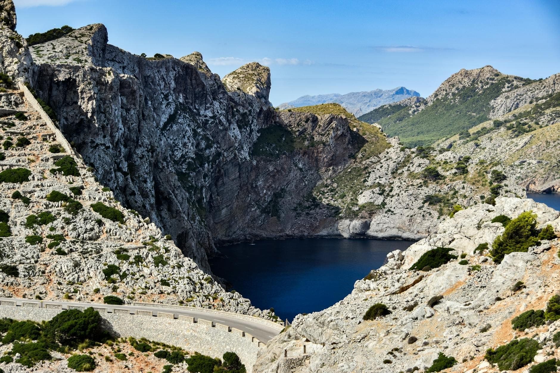 Una tortuosa strada di montagna abbraccia scogliere rocciose sopra una spettacolare baia, con le vette della Serra de Tramuntana sullo sfondo a Maiorca.