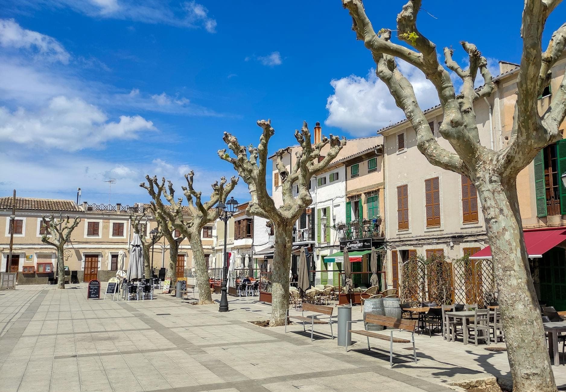 Praça aberta de aldeia em Mallorca com árvores podadas, edifícios de pedra e cafés ao ar livre sob um céu azul brilhante.