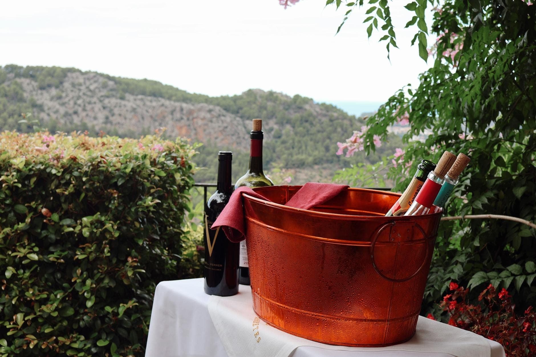 Mesa ao ar livre com balde de gelo de cobre segurando garrafas de vinho, cercada por vegetação exuberante e vistas panorâmicas das montanhas, evocando o clima da região vinícola de Maiorca.