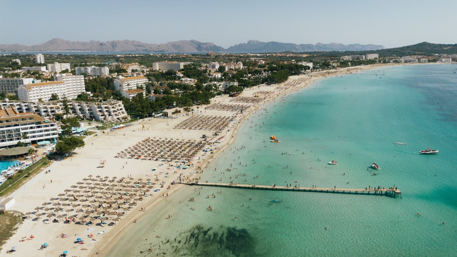 Luftaufnahme eines großen Sandstrands auf Mallorca mit Liegestühlen, Sonnenschirmen, türkisfarbenem Wasser und nahegelegenen Hotels, ideal für Familien.