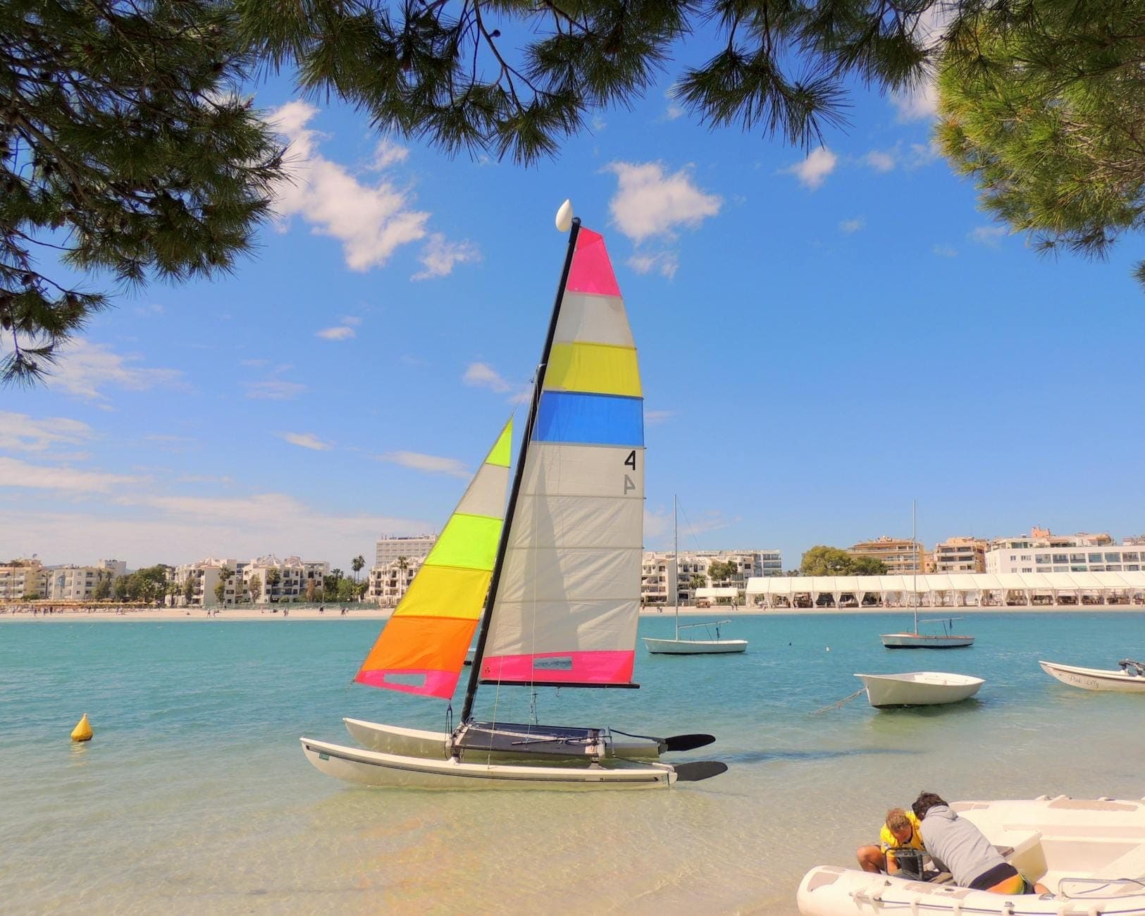 Buntes Segelboot an einem ruhigen Strand auf Mallorca, türkisblaues klares Wasser, Kinder spielen am Ufer, strahlend blauer Himmel mit vereinzelten Wolken.