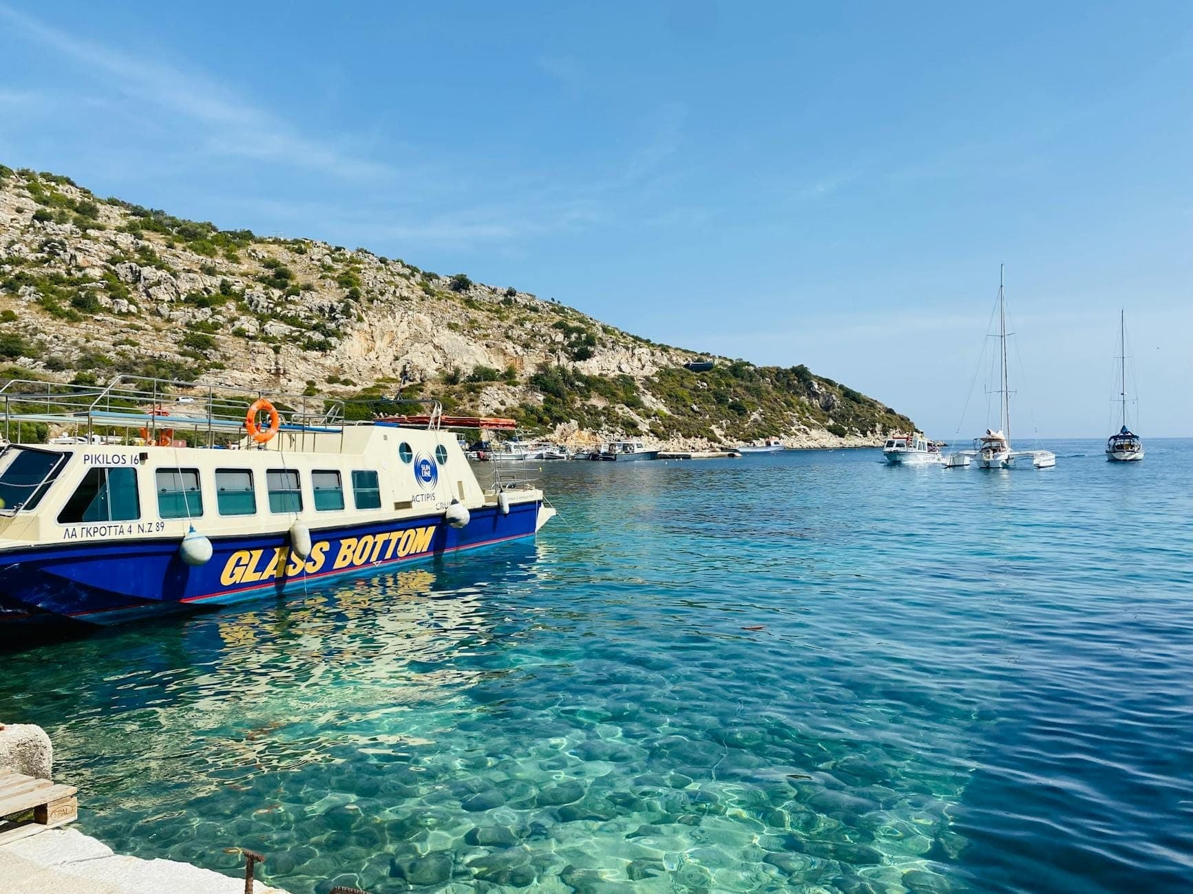 Ein Glasbodenboot, das in der Nähe des Ufers angelegt hat, mit klarem türkisfarbenem Wasser, felsigen Hügeln und Segelbooten im Hintergrund auf Mallorca.