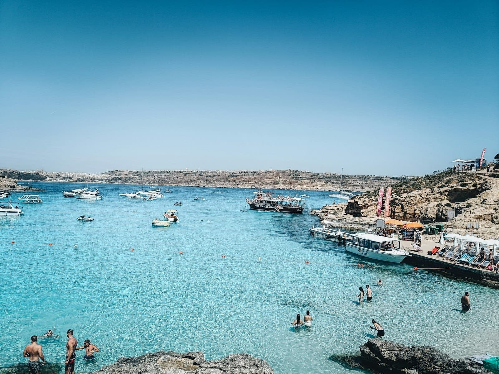 Klares türkisfarbenes Wasser der Blue Lagoon mit Schwimmern, felsigem Ufer und verankerten Booten in Comino, Malta, unter einem strahlend blauen Himmel.