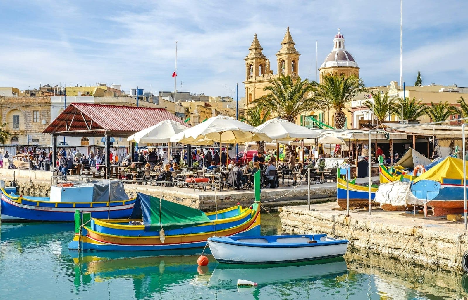 Bunte traditionelle Fischerboote im Hafen von Marsaxlokk mit Marktständen, Sonnenschirmen und der Kirche im Hintergrund unter blauem Himmel.