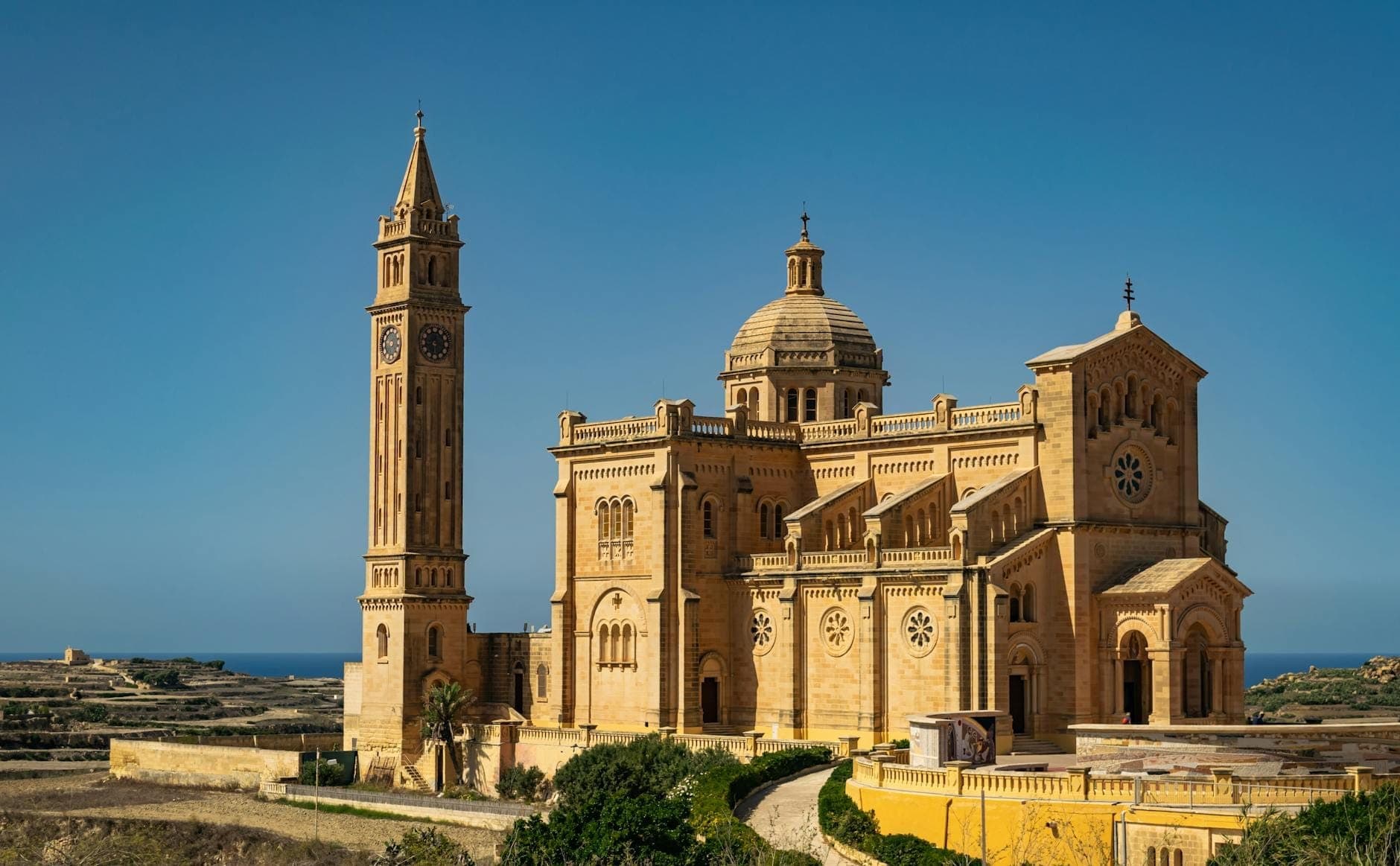Beeindruckende Sandsteinkirche mit einem hohen Uhrenturm in der ländlichen Landschaft von Gozo unter klarem blauem Himmel.
