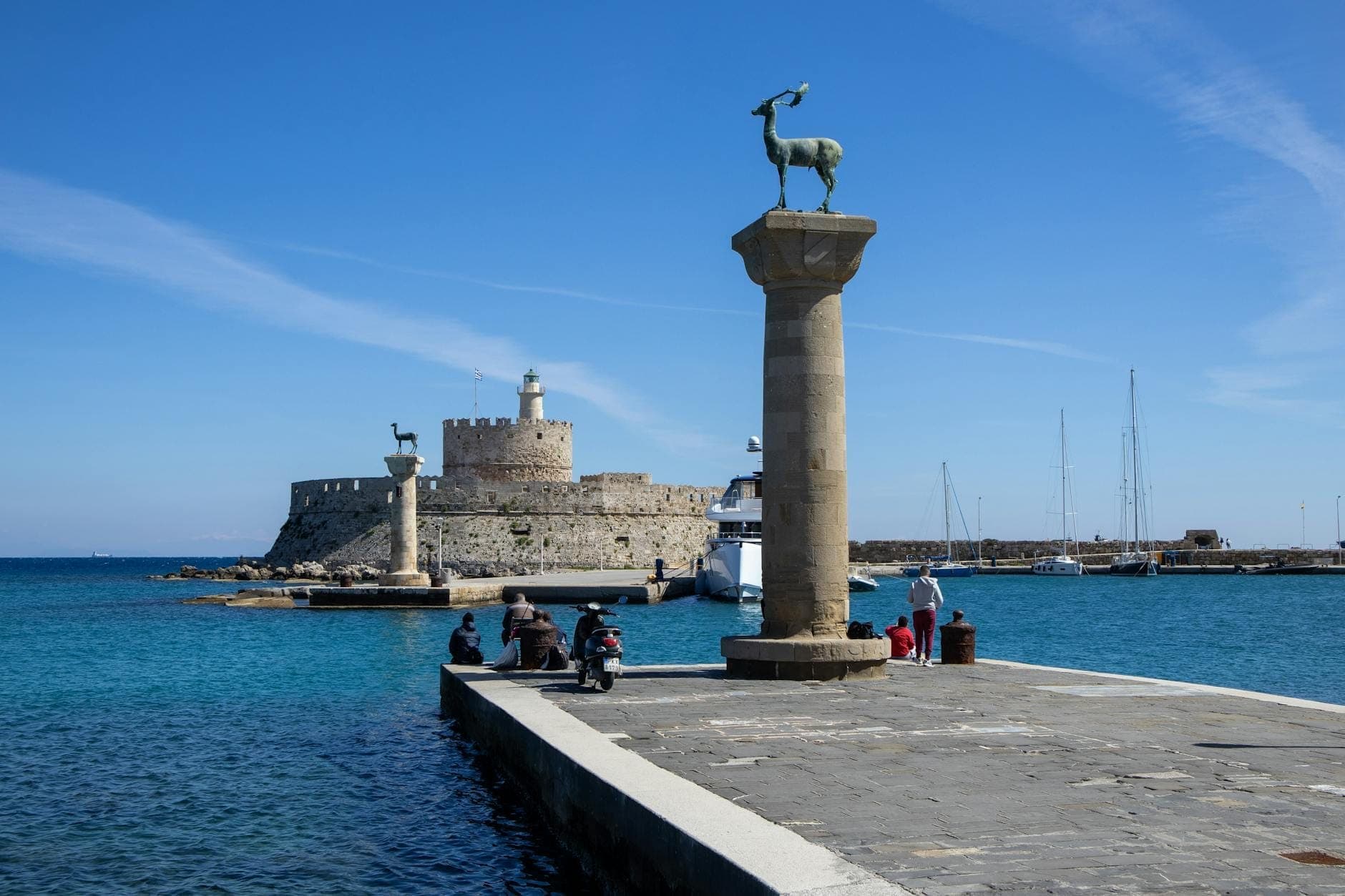 Entrée du port de Mandraki avec ses statues de cerfs en bronze sur leurs colonnes, le fort Saint-Nicolas en arrière-plan, un ciel bleu limpide et des bateaux amarrés dans les eaux turquoise.