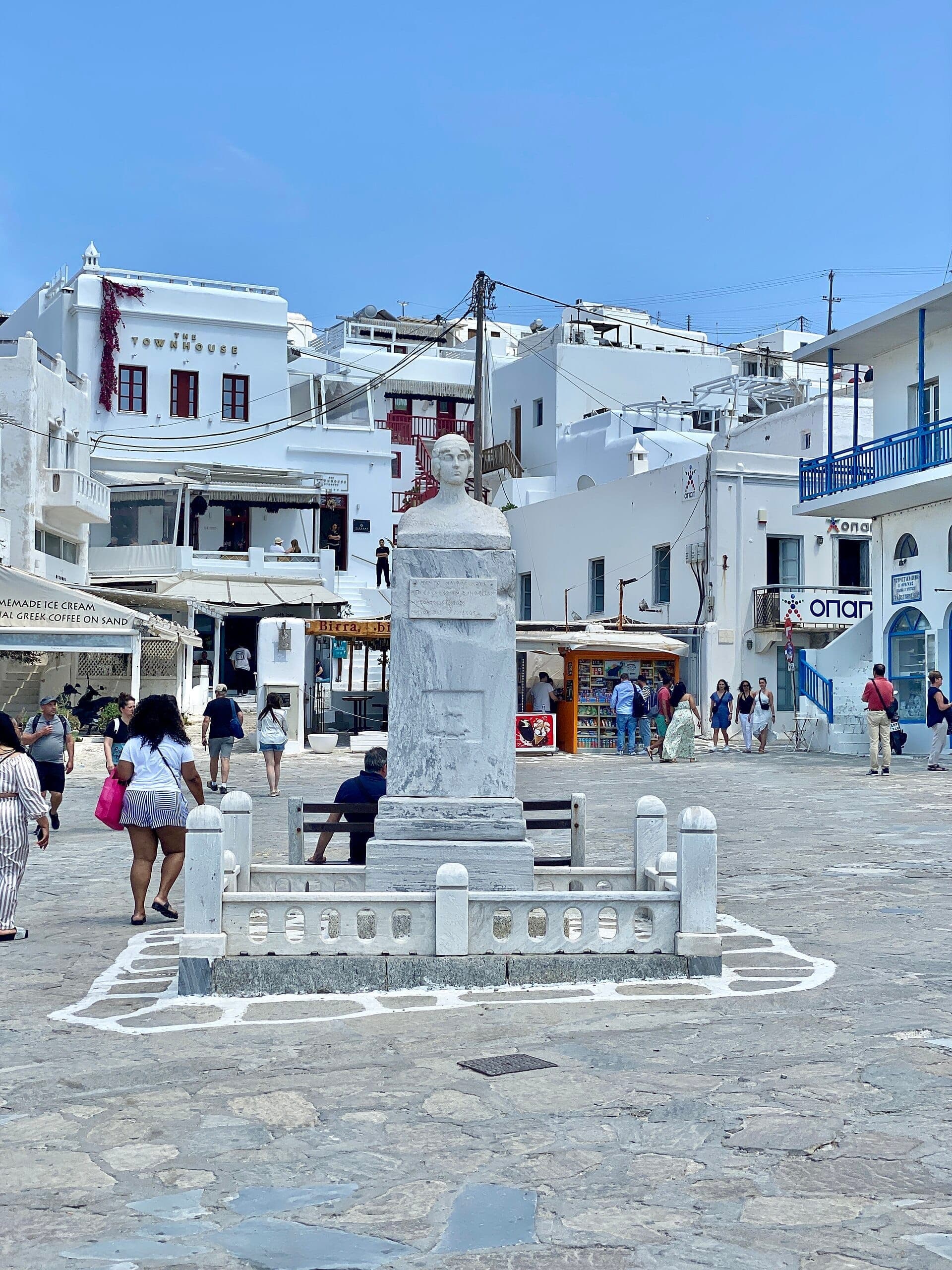 Marmorstatue von Manto Mavrogenous im Zentrum des Platzes, umgeben von weißen kykladischen Gebäuden und Menschen, die Mykonos Town erkunden.