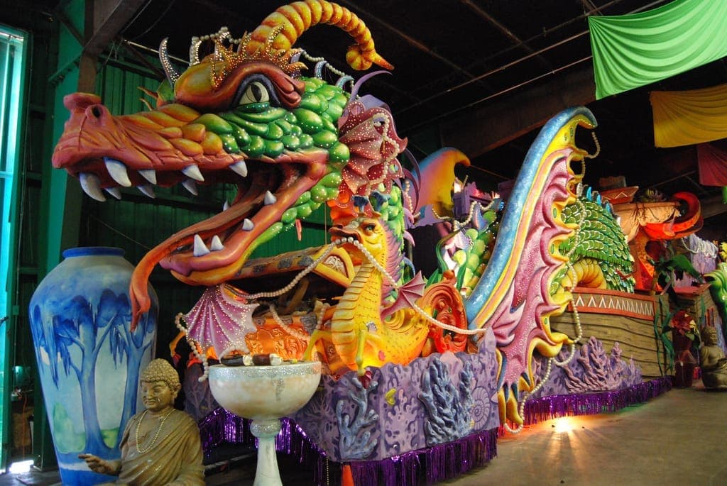 Giant, colorful dragon-themed parade float under construction at Mardi Gras World, surrounded by vibrant decorations and sculptures inside a spacious warehouse.