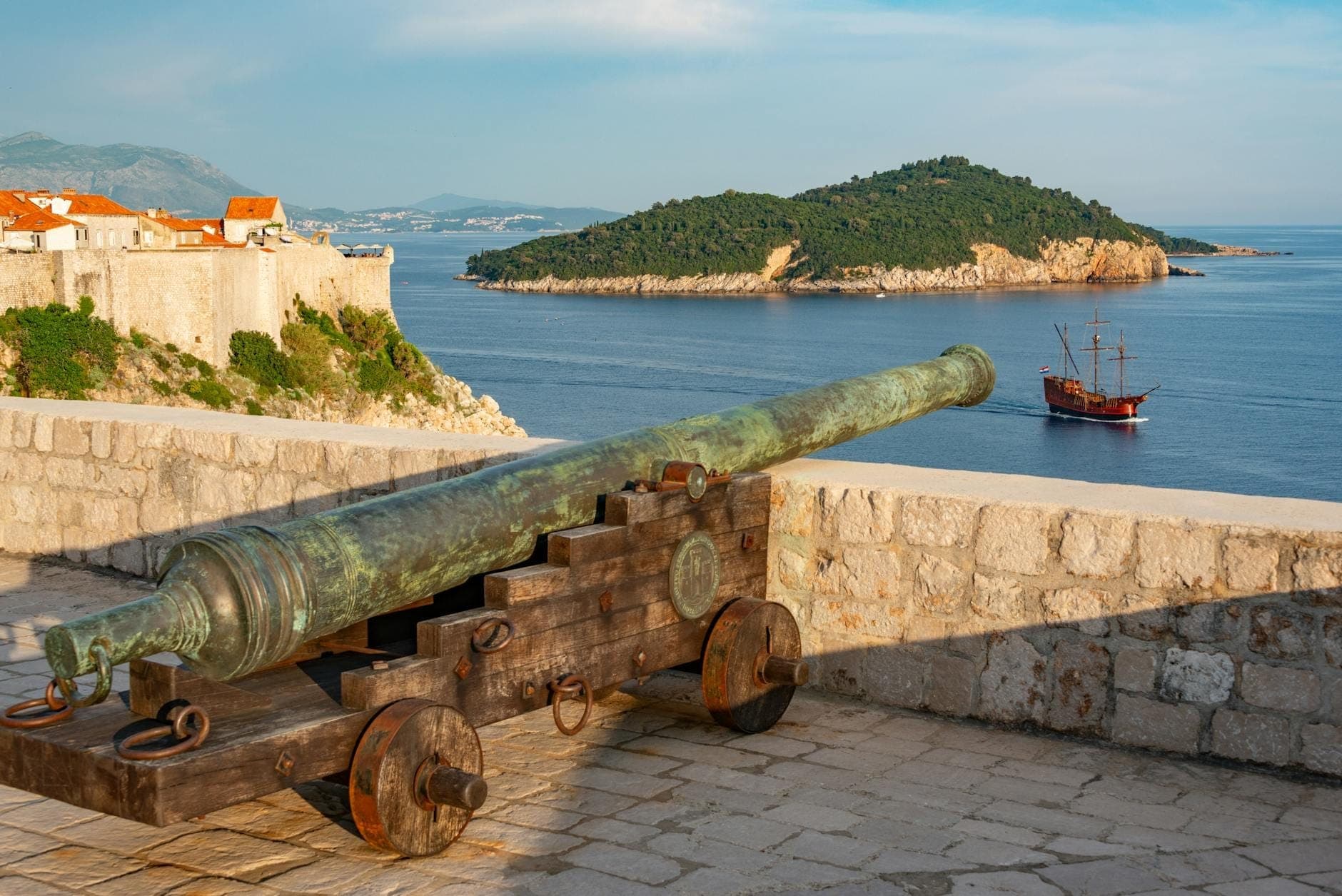 Eine alte Bronzekanone auf der Steinmauer der Dubrovniker Festung mit Blick auf die blaue Adria, Befestigungsanlagen, eine grüne Insel und ein Segelschiff in der Ferne.