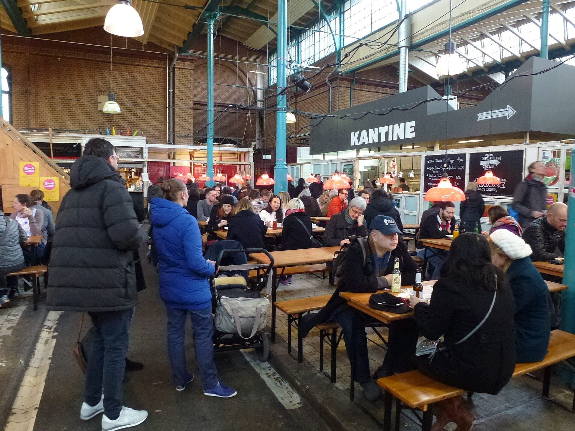 นักท่องเที่ยวนั่งรวมตัวกันที่โต๊ะไม้ยาวภายใน Markthalle Neun ท่ามกลางเพดานสูง ผนังอิฐโชว์ และร้านอาหารที่คึกคักเป็นฉากหลัง