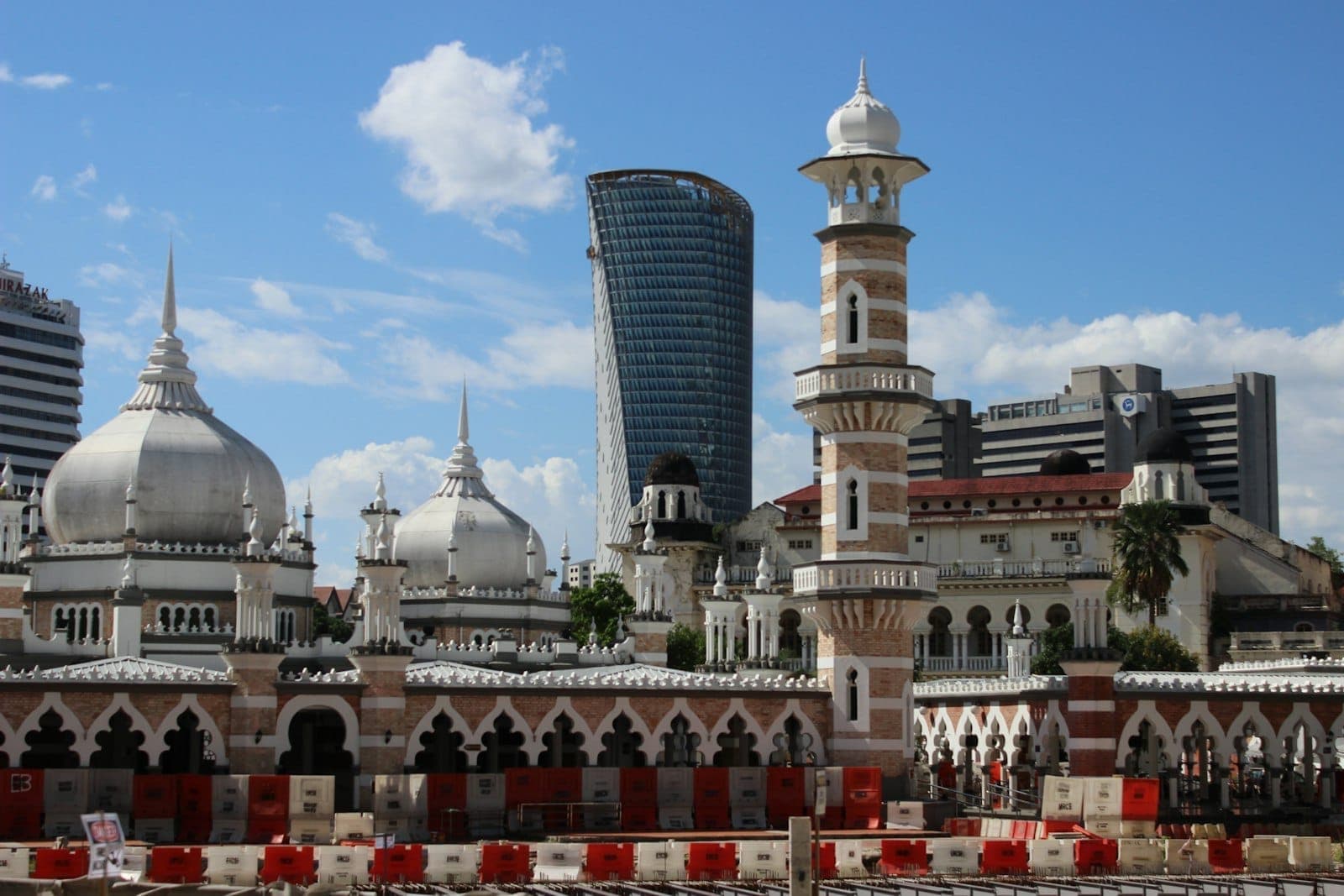 Masjid Jamek'in Babürlü tarzı kubbelerine ve uzun minaresine mavi gökyüzünün altında, arka planda modern Kuala Lumpur gökdelenleriyle birlikte bakılmaktadır.