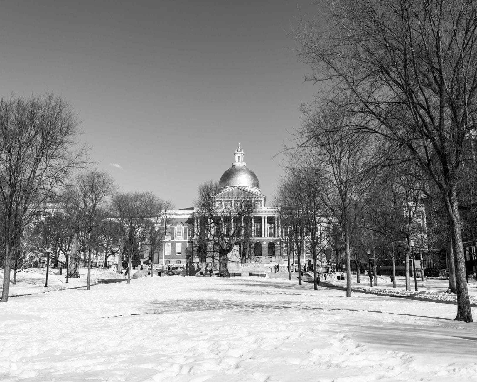 Scène hivernale en noir et blanc du Massachusetts State House avec son emblématique dôme doré, encadré par le Boston Common enneigé et ses arbres dénudés.