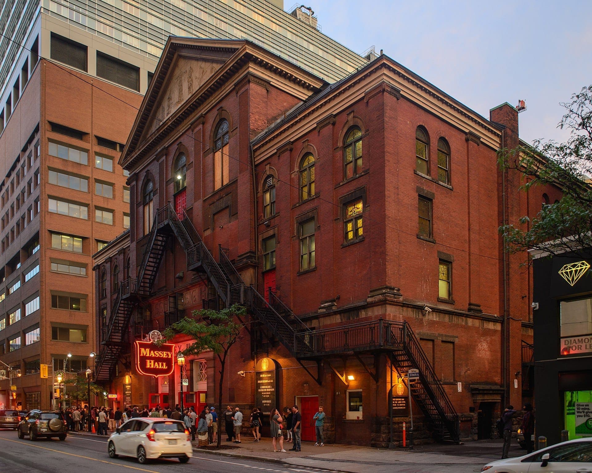 La storica facciata in mattoni rossi della Massey Hall al tramonto, con un'insegna al neon e persone in fila sul marciapiede nel centro di Toronto.