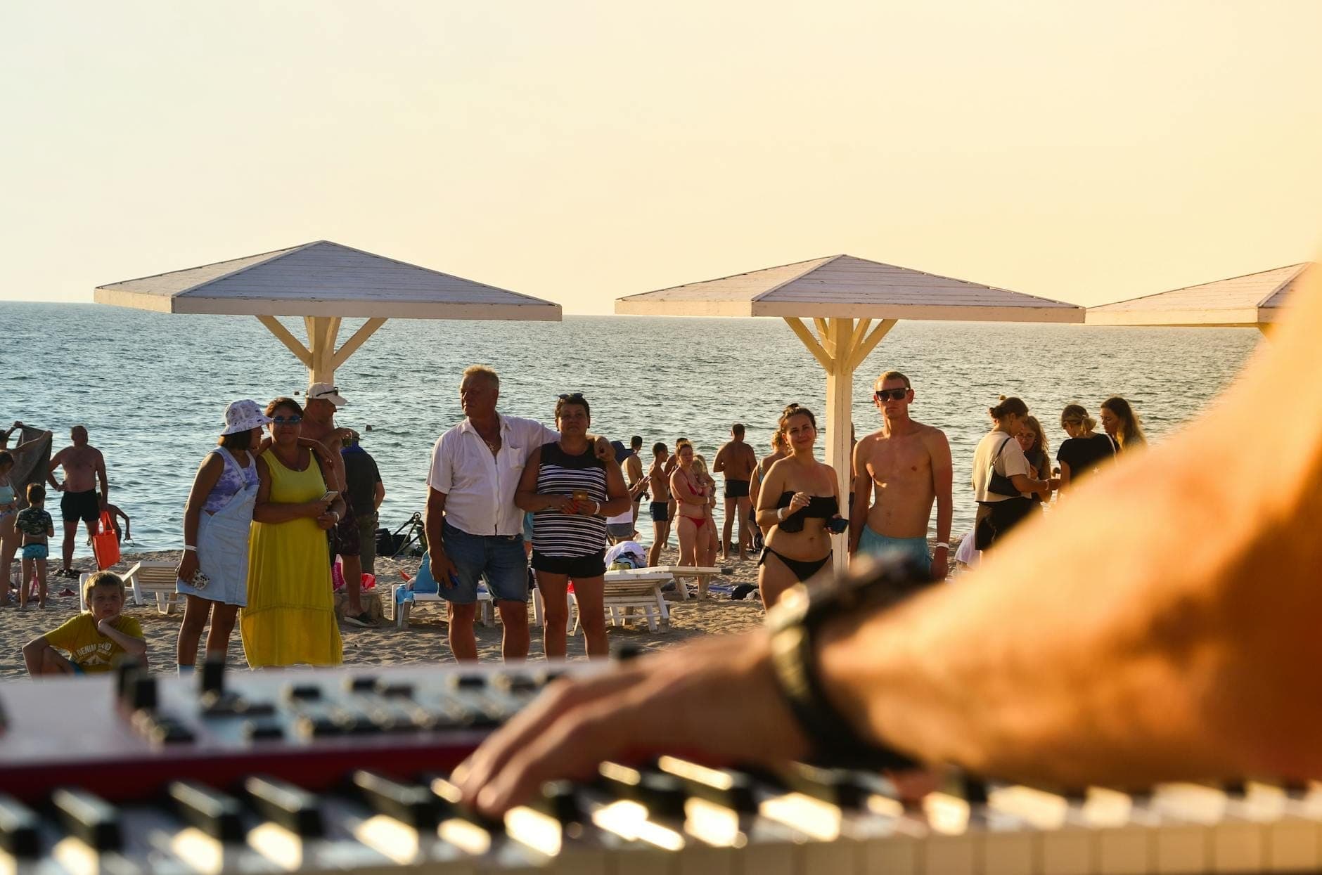 Nahaufnahme eines Keyboardspielers an einem belebten Strand mit versammelten Menschen, Sonnenschirmen und dem Meer im Hintergrund bei Tageslicht.