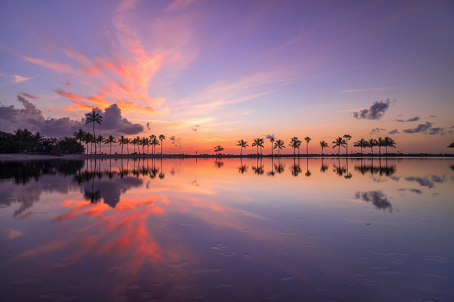 View of Matheson Hammock Park Beach at sunset with palm trees reflected in calm, shallow water under a vibrant purple and orange sky.
