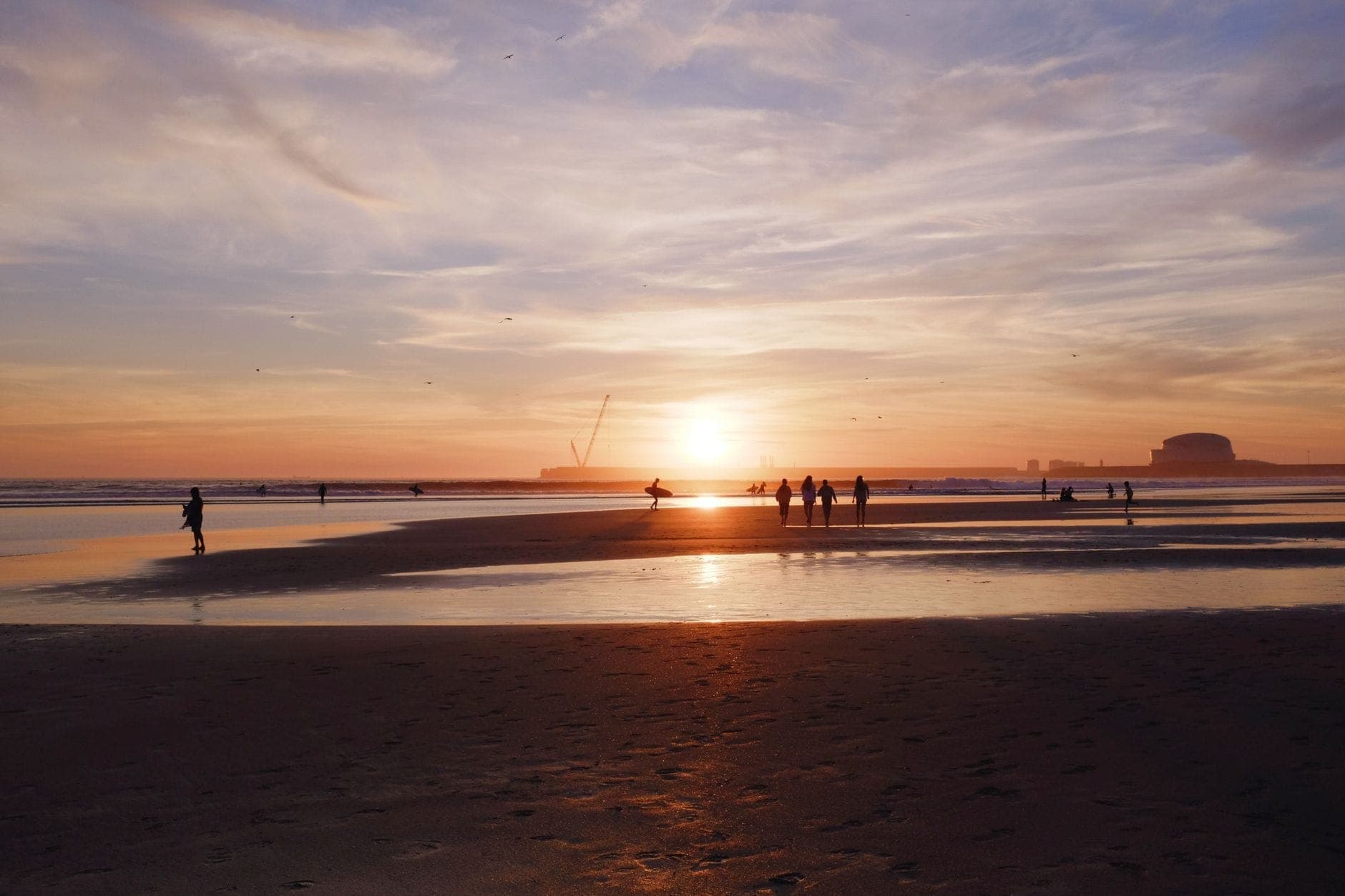 Gruppen von Menschen genießen einen goldenen Sonnenuntergang auf dem weitläufigen Sandstrand von Matosinhos mit den Atlantikwellen und den Hafenanlagen im Hintergrund.