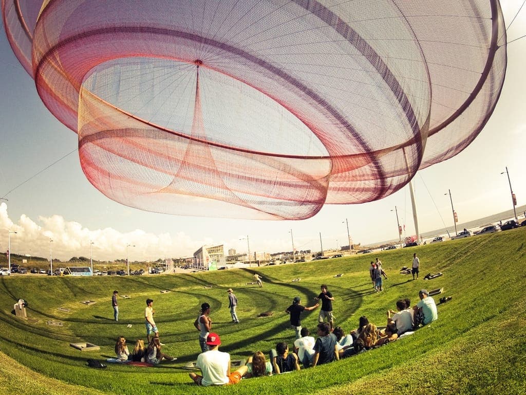Große zeitgenössische Netzskulptur, die über einem begrünten öffentlichen Park in Matosinhos hängt, mit Gruppen von Menschen, die an einem sonnigen Tag darunter entspannen.