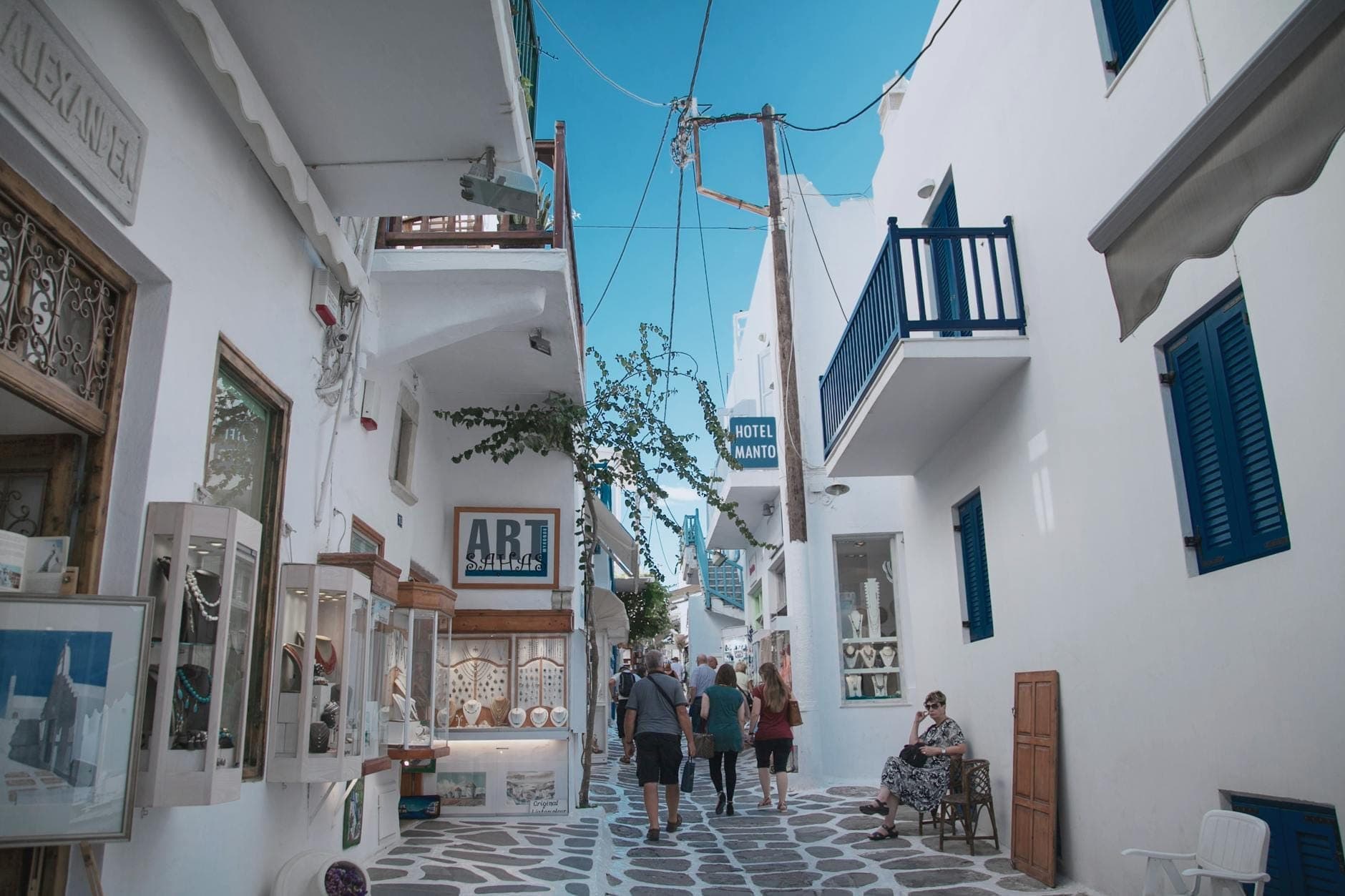 Menschen schlendern durch die Matoyianni Street in Mykonos Town, umgeben von weißen zykladischen Gebäuden, Boutiquen und Cafés unter einem strahlend blauen Himmel.