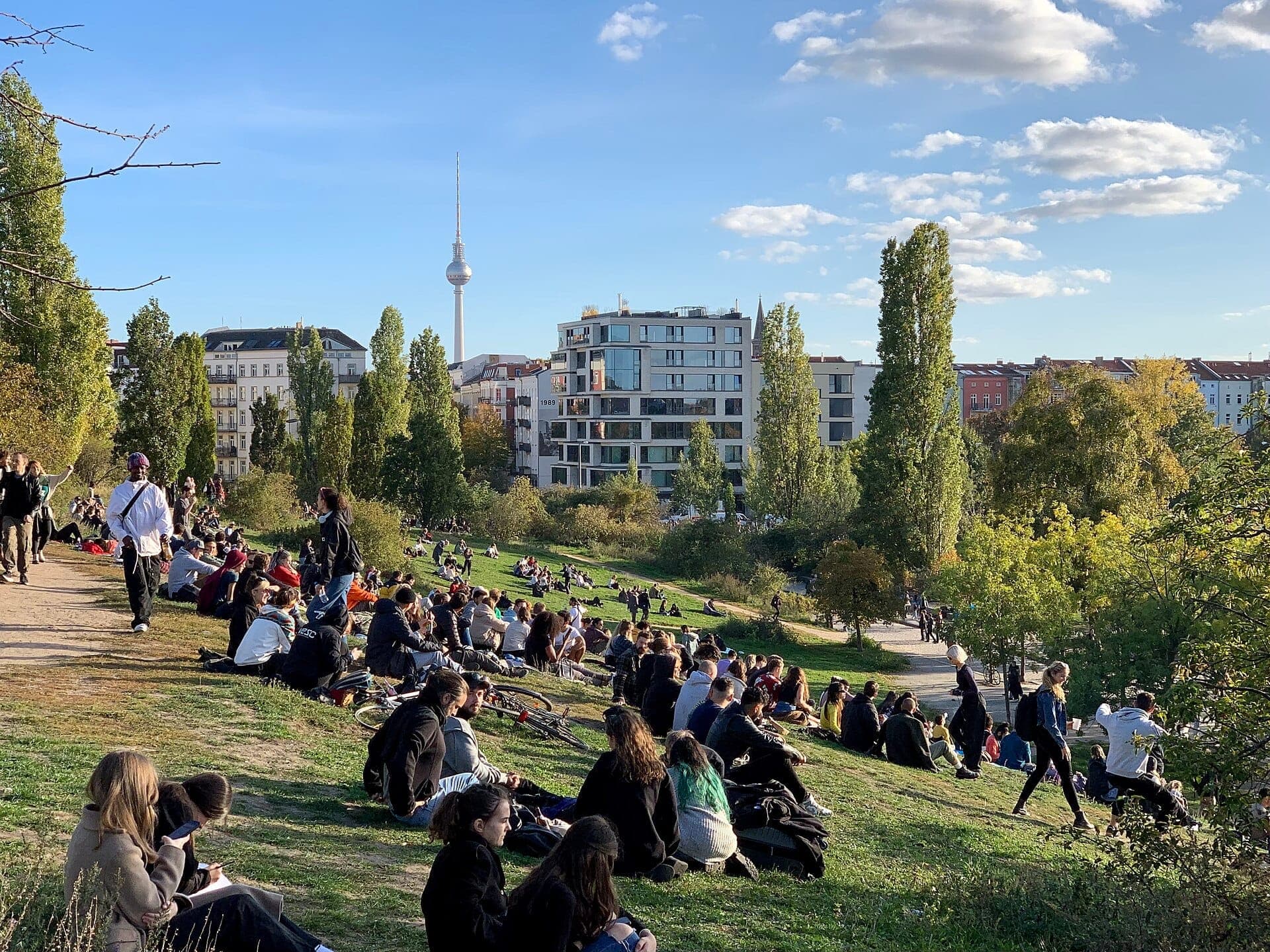 ฝูงชนผ่อนคลายและพบปะสังสรรค์บนเนินหญ้าใน Mauerpark กรุงเบอร์ลิน โดยมีอาคารในเมืองและหอโทรทัศน์ปรากฏให้เห็นใต้ท้องฟ้าสีฟ้า