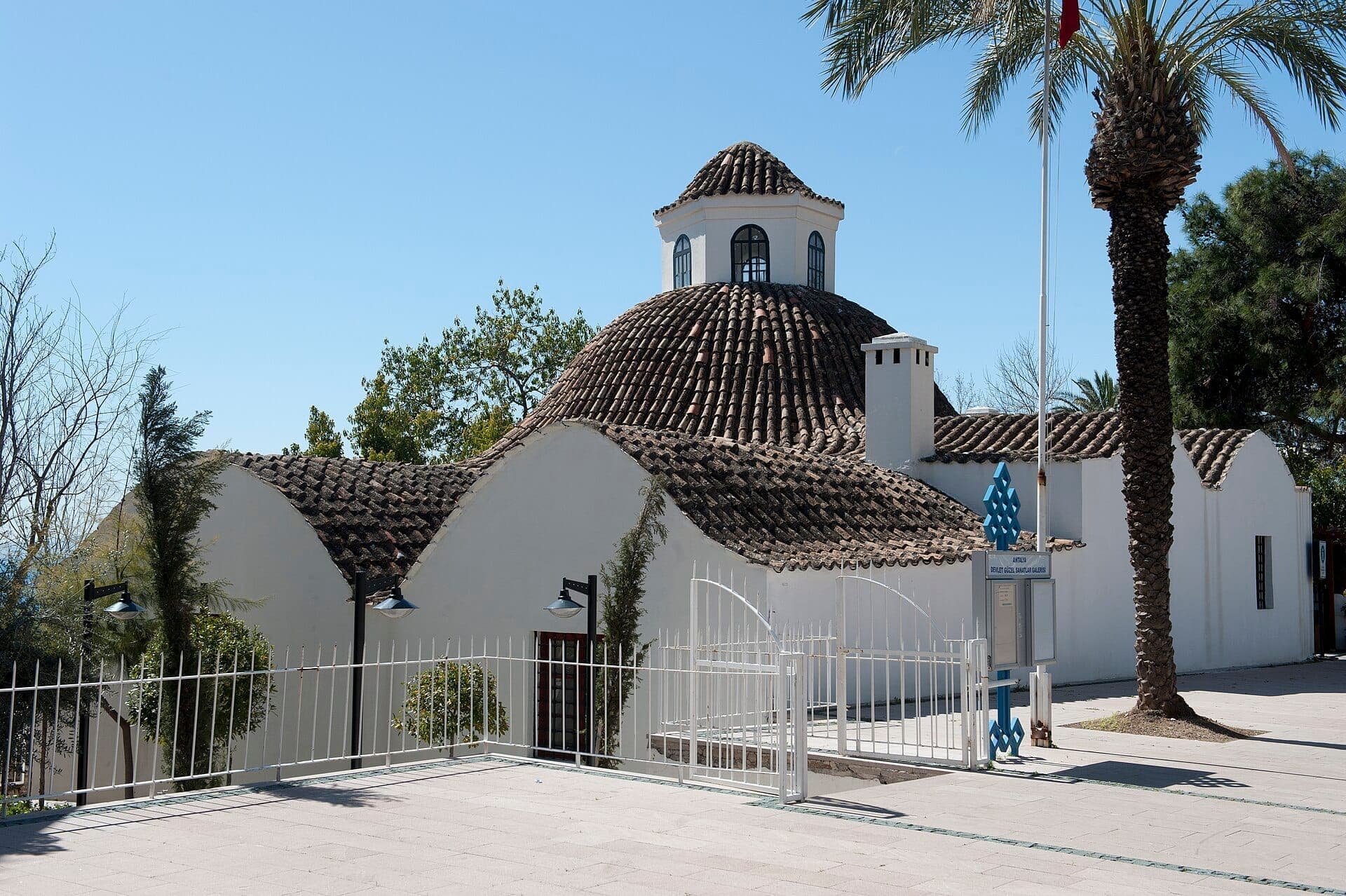 Das Mevlevi-Klostermuseum Antalya zeigt weiß getünchte Wände und ein traditionelles Kuppeldach, eingerahmt von Palmen und in Tageslicht getaucht.
