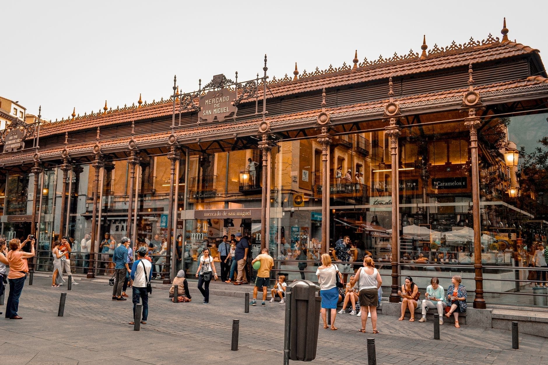 Широкий вид на нарядный фасад Mercado de San Miguel из стекла и чугуна с оживлёнными людьми снаружи — атмосфера в самом центре Мадрида.