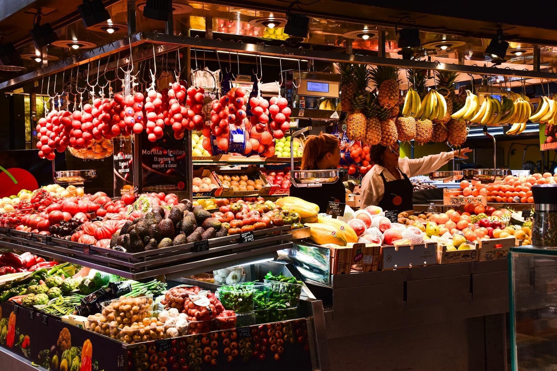Bunter Obst- und Gemüsestand im Mercat de Santa Catalina mit Tomaten, Bananen, Ananas und Einkäufern unter warmem Innenlicht.