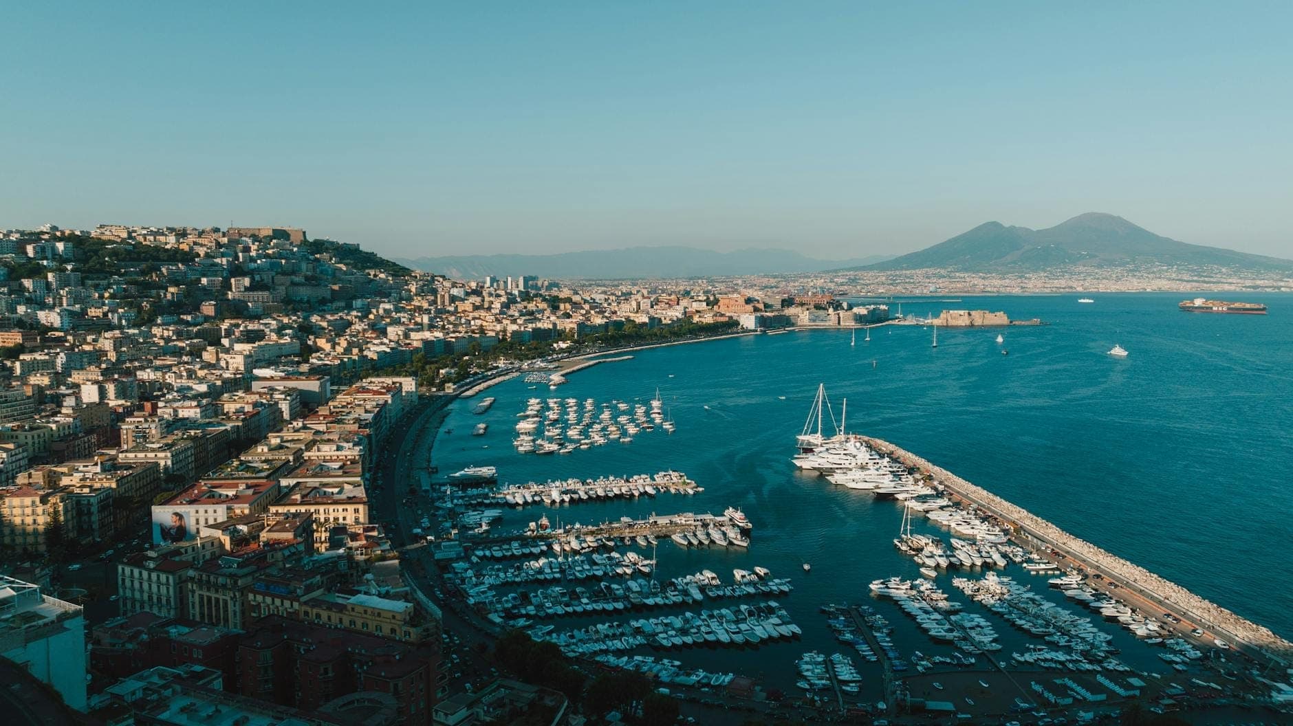 Vista aérea do Porto de Mergellina com barcos atracados na marina, orla de Nápoles, prédios da cidade e o Monte Vesúvio ao fundo sob um céu azul claro.