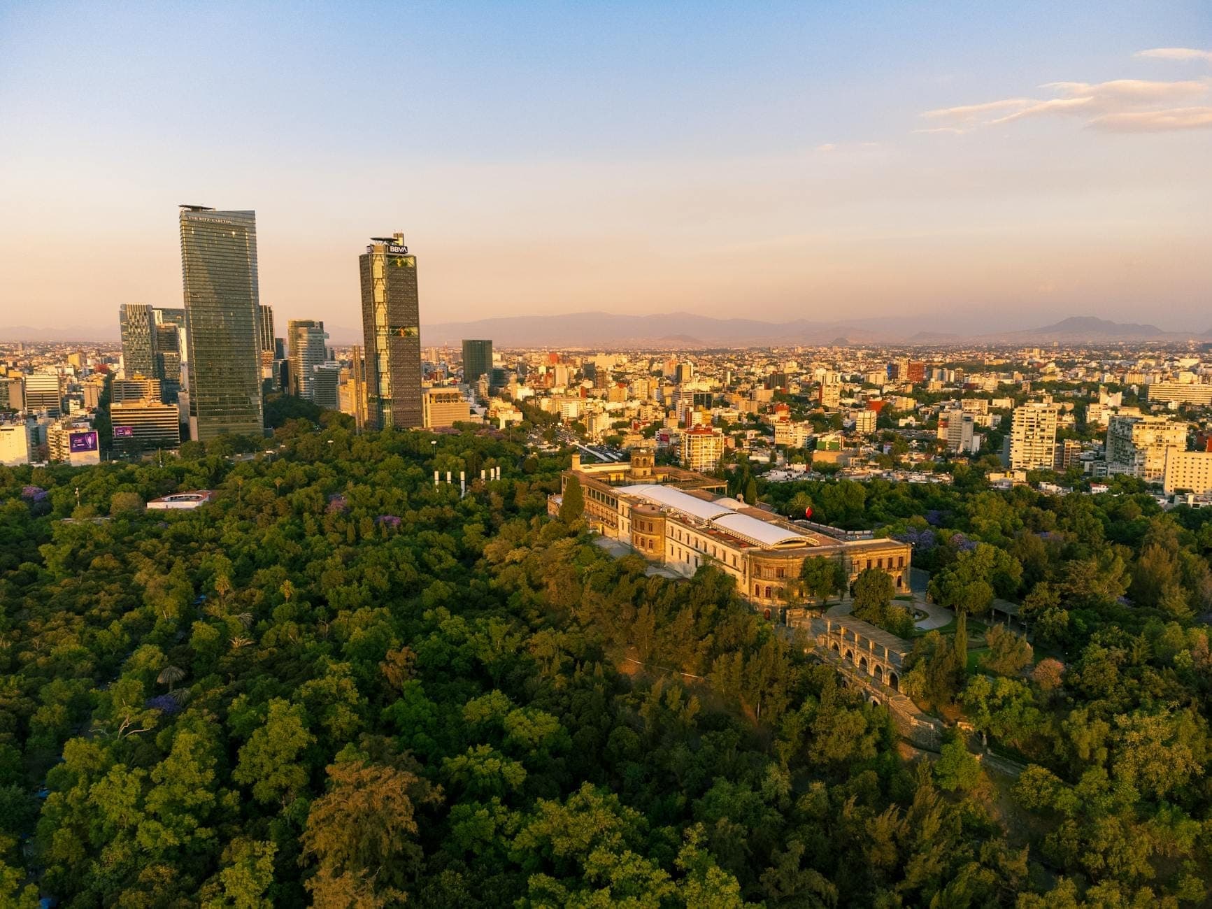 Aerial view of Chapultepec Castle atop a lush green hill surrounded by forest, with Mexico City skyline in the background at sunset.