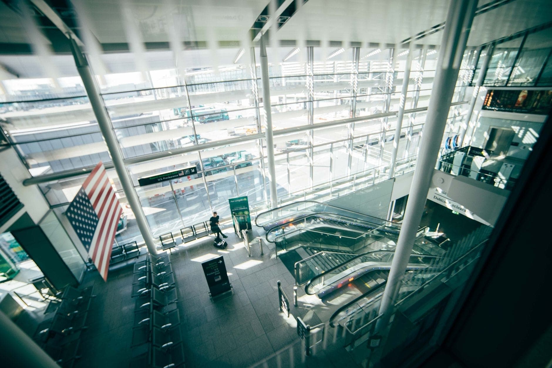 Bright and modern airport terminal interior with large windows, escalators, and a prominent American flag, showing a spacious arrivals area in daylight.
