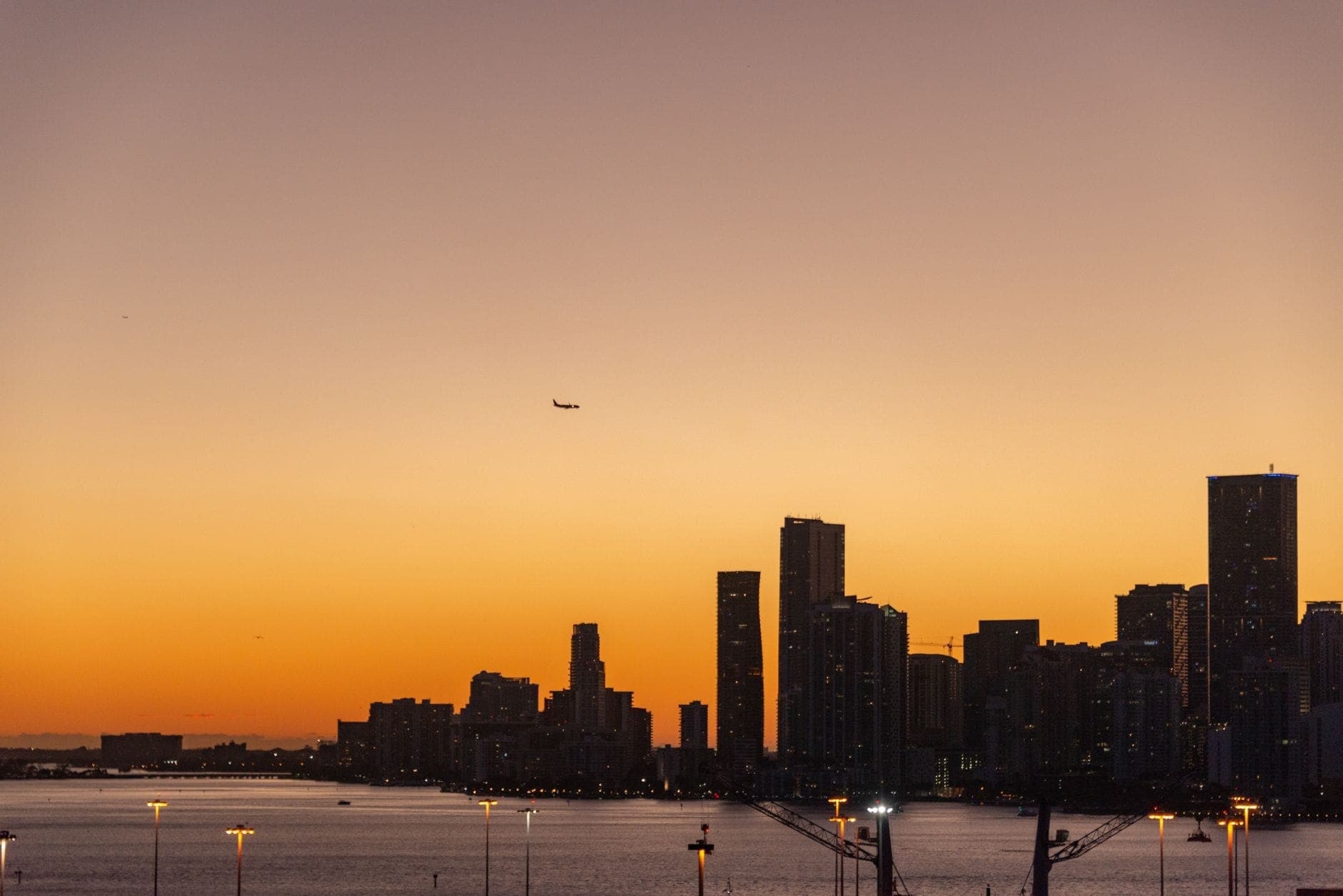 Miami skyline at sunset with a plane approaching above the city and water in the foreground
