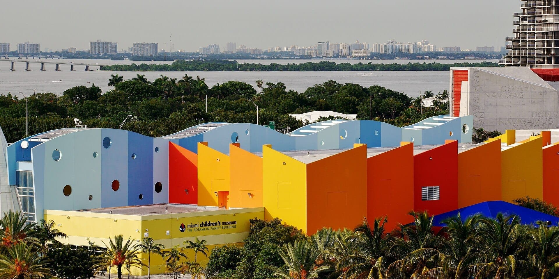 Exterior view of Miami Children's Museum featuring colorful geometric facades, palm trees, and Biscayne Bay with Miami skyline in the background.