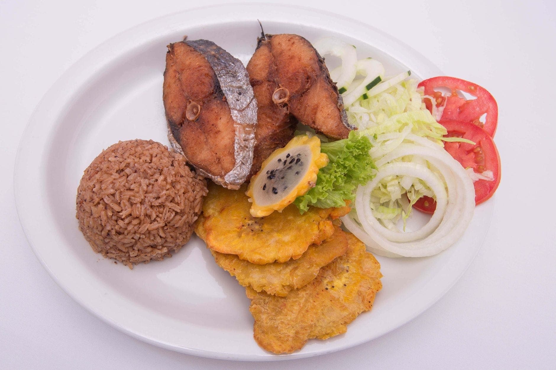 A plate with classic Cuban food: fried fish, tostones (fried plantains), rice with beans, lettuce, onions, and tomato, served on a white background.