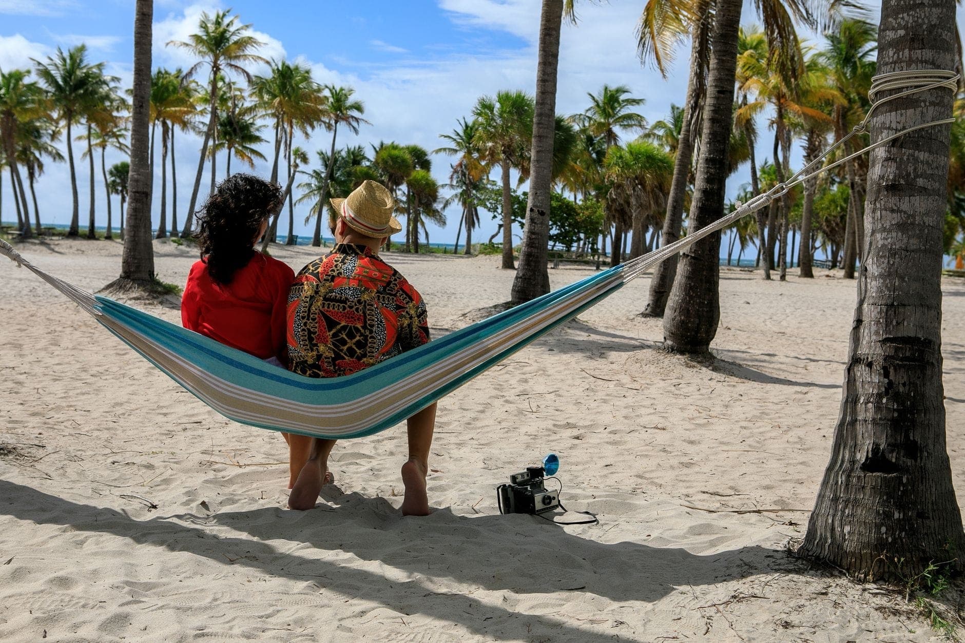 Couple relaxing together in a hammock strung between palm trees on a sandy Miami beach, with lush palms and blue sky in the background.