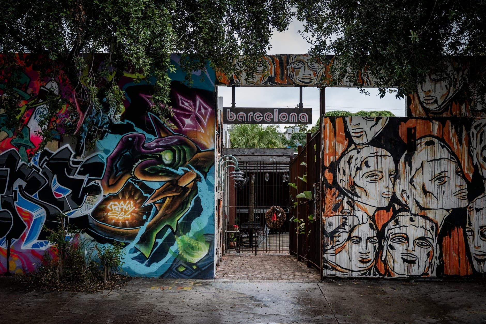 Entrance to a hidden Miami courtyard flanked by brightly colored street art and murals, with lush trees arching overhead and a glimpse of seating inside.