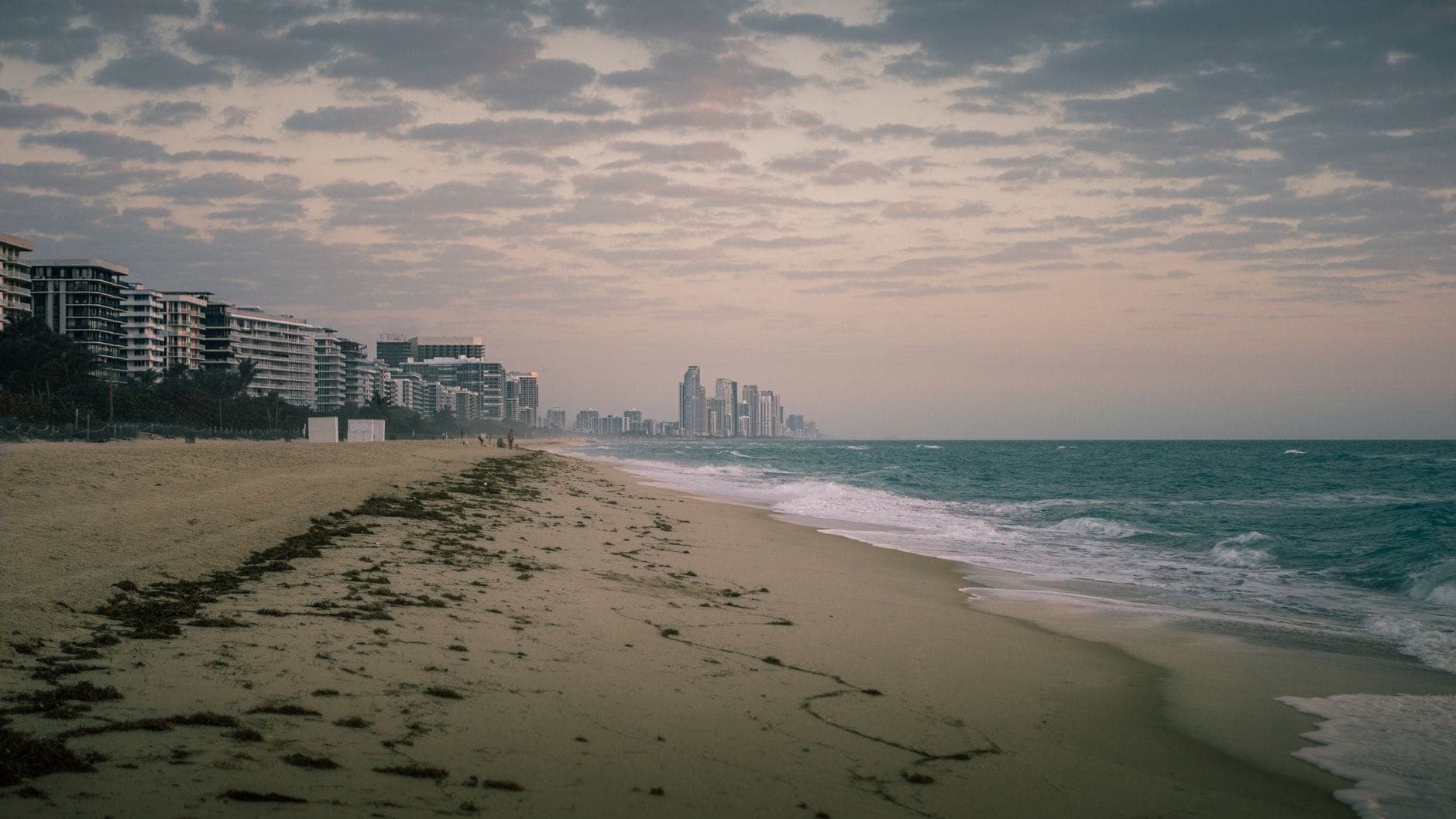 Wide view of Miami Beach shoreline in winter, with modern high-rise buildings, overcast skies, and gentle waves along the sandy coast.