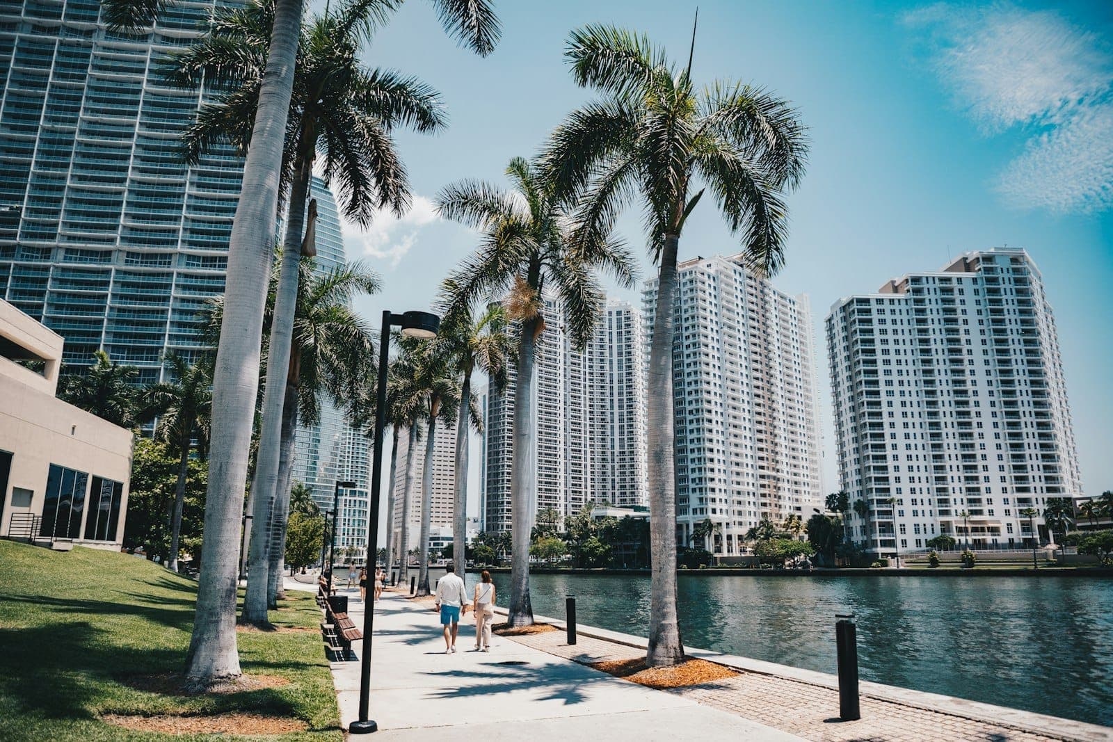 People walking along the palm tree-lined Miami Riverwalk with modern high-rise buildings and the river visible on a sunny day.