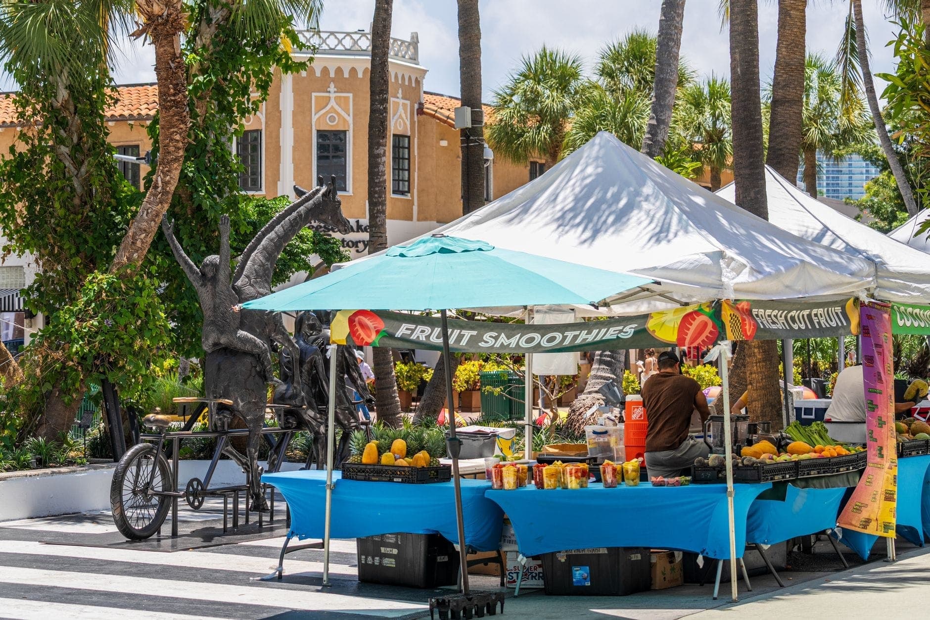 Scène de marché en plein air à Miami avec des étals de fruits sous des tentes, des palmiers et des bâtiments de style méditerranéen en arrière-plan.