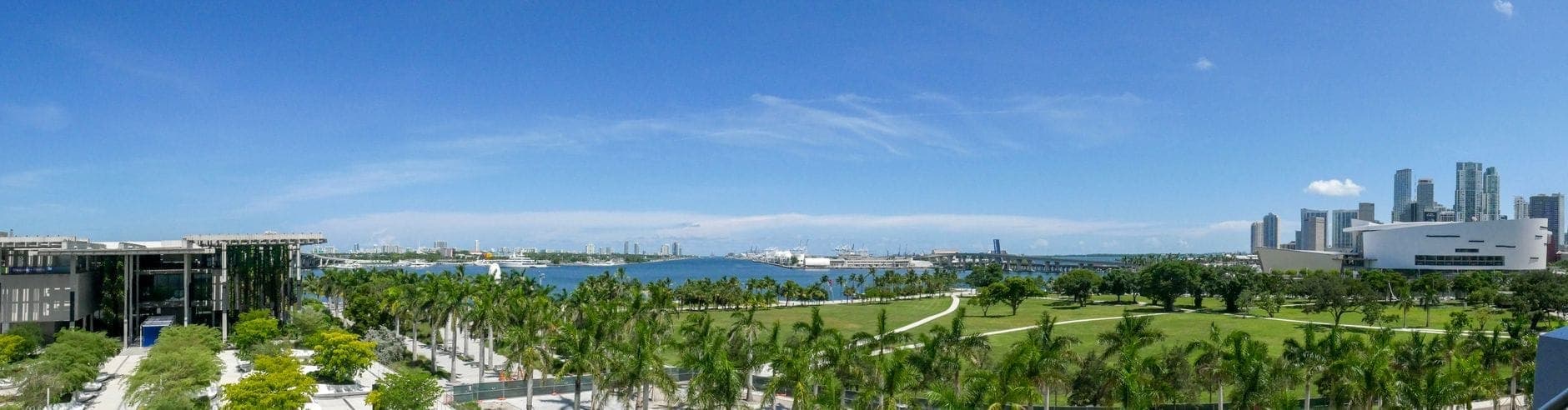 Panoramic view of Miami skyline with palm trees, blue sky, and Biscayne Bay, showing both cityscape and tropical weather typical of Miami.