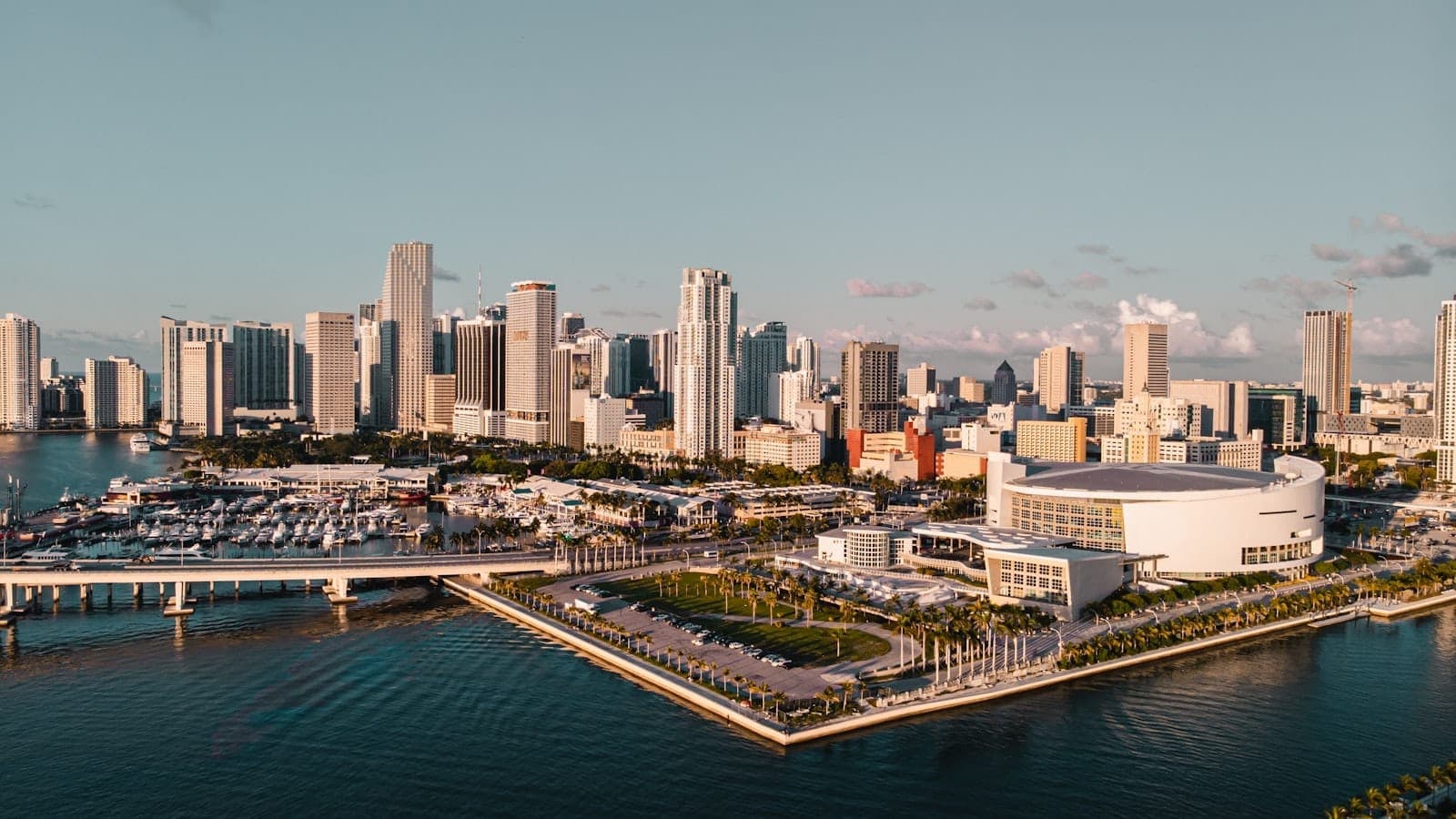 A wide, sweeping aerial view of downtown Miami featuring iconic skyscrapers, the bay, marina, and the FTX Arena in golden evening sunlight.