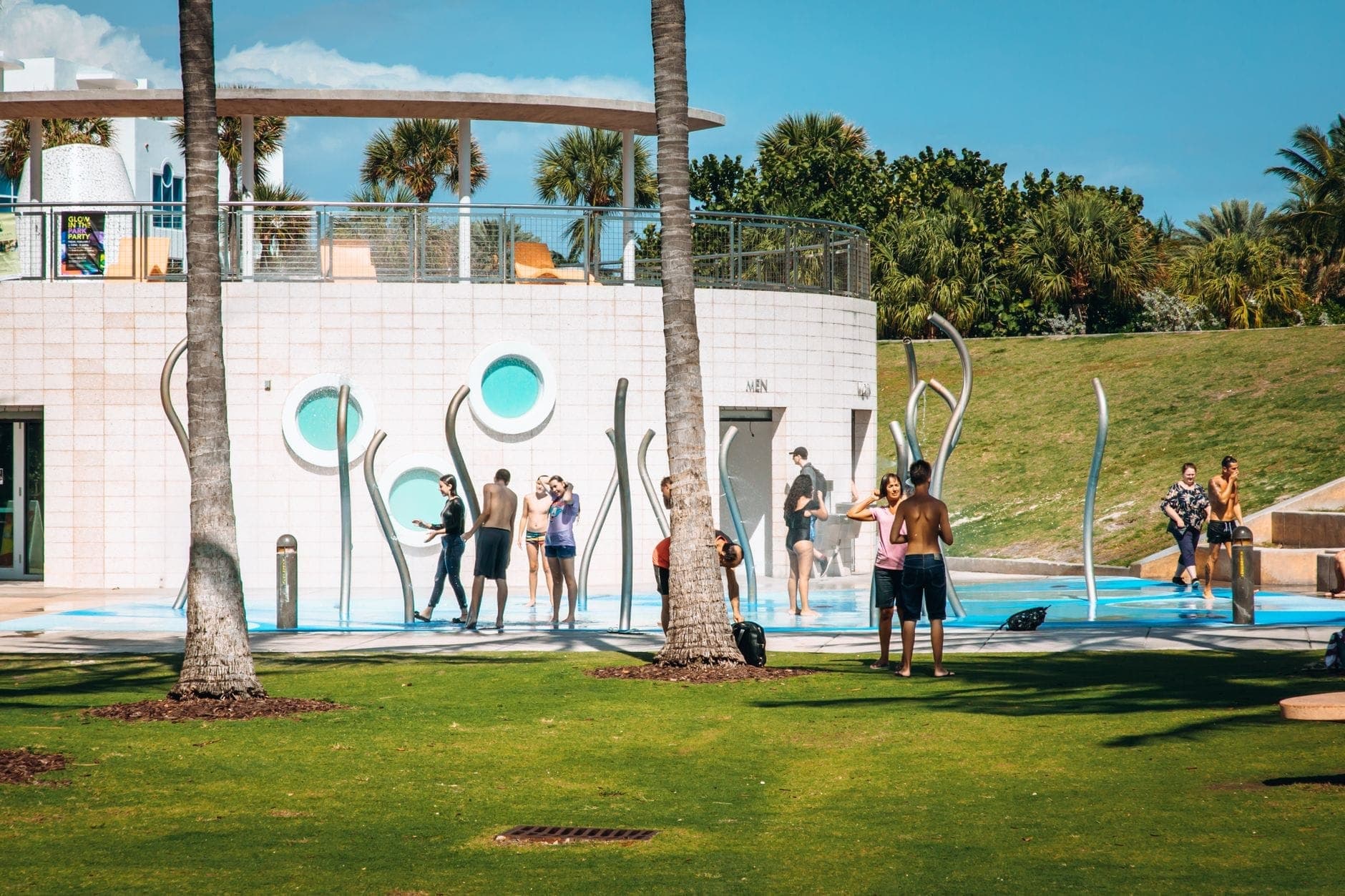 Children and families play in a colorful splash pad area surrounded by palm trees in a sunny Miami park, perfect for kids and families.