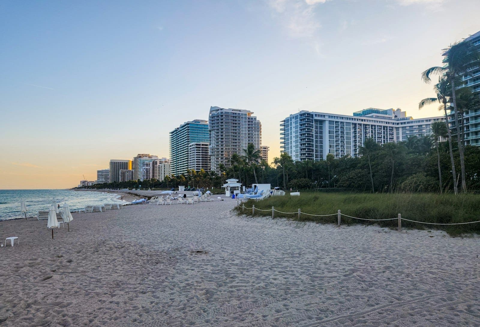 View of a wide sandy beach lined with modern resort hotels, palm trees, and calm ocean waters under a clear sky in Miami Beach.