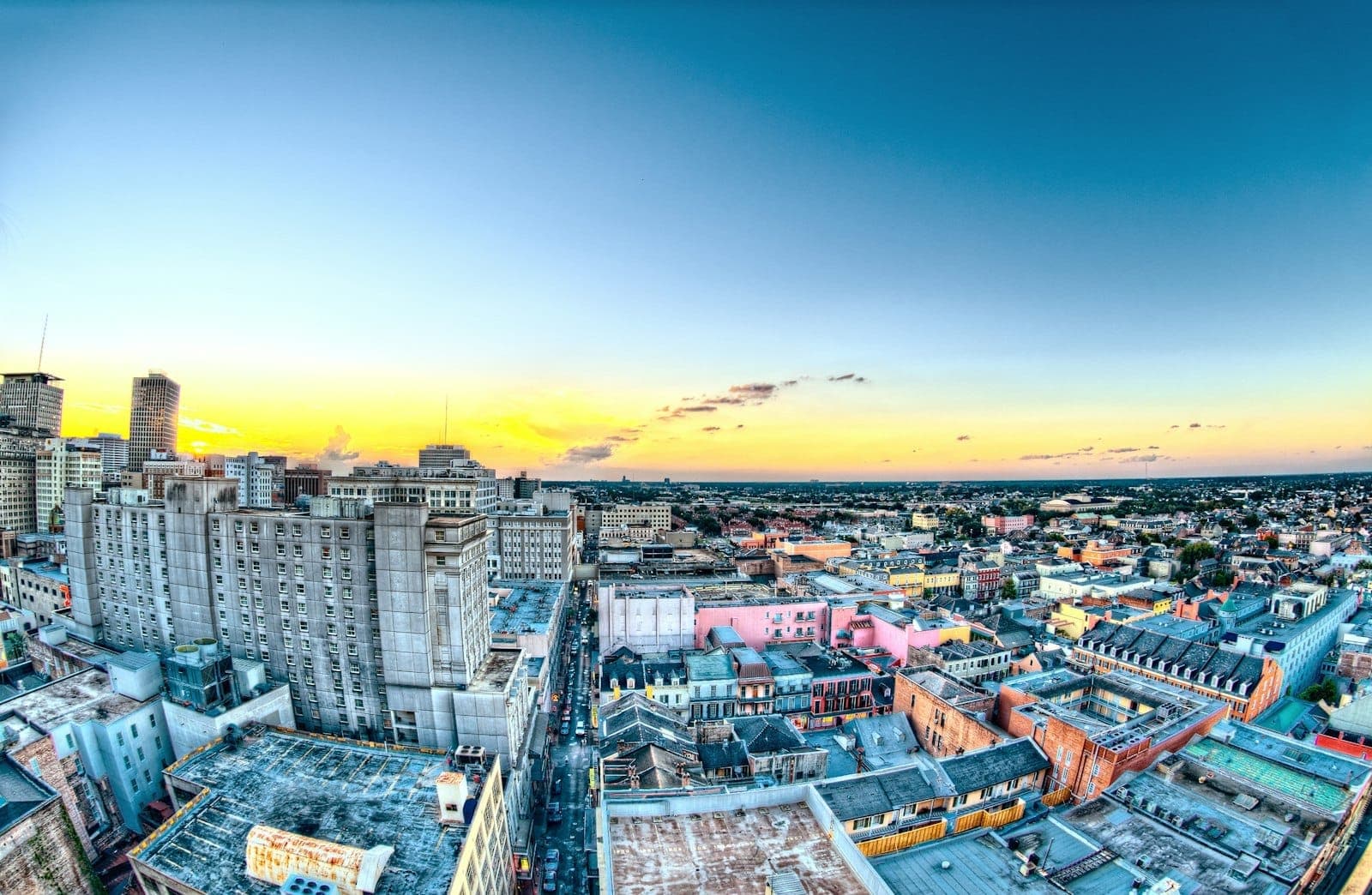 Colorful, wide-angle aerial view of New Orleans’ Mid-City at sunrise, with city buildings, rooftops, and a glowing blue and yellow sky in the background.